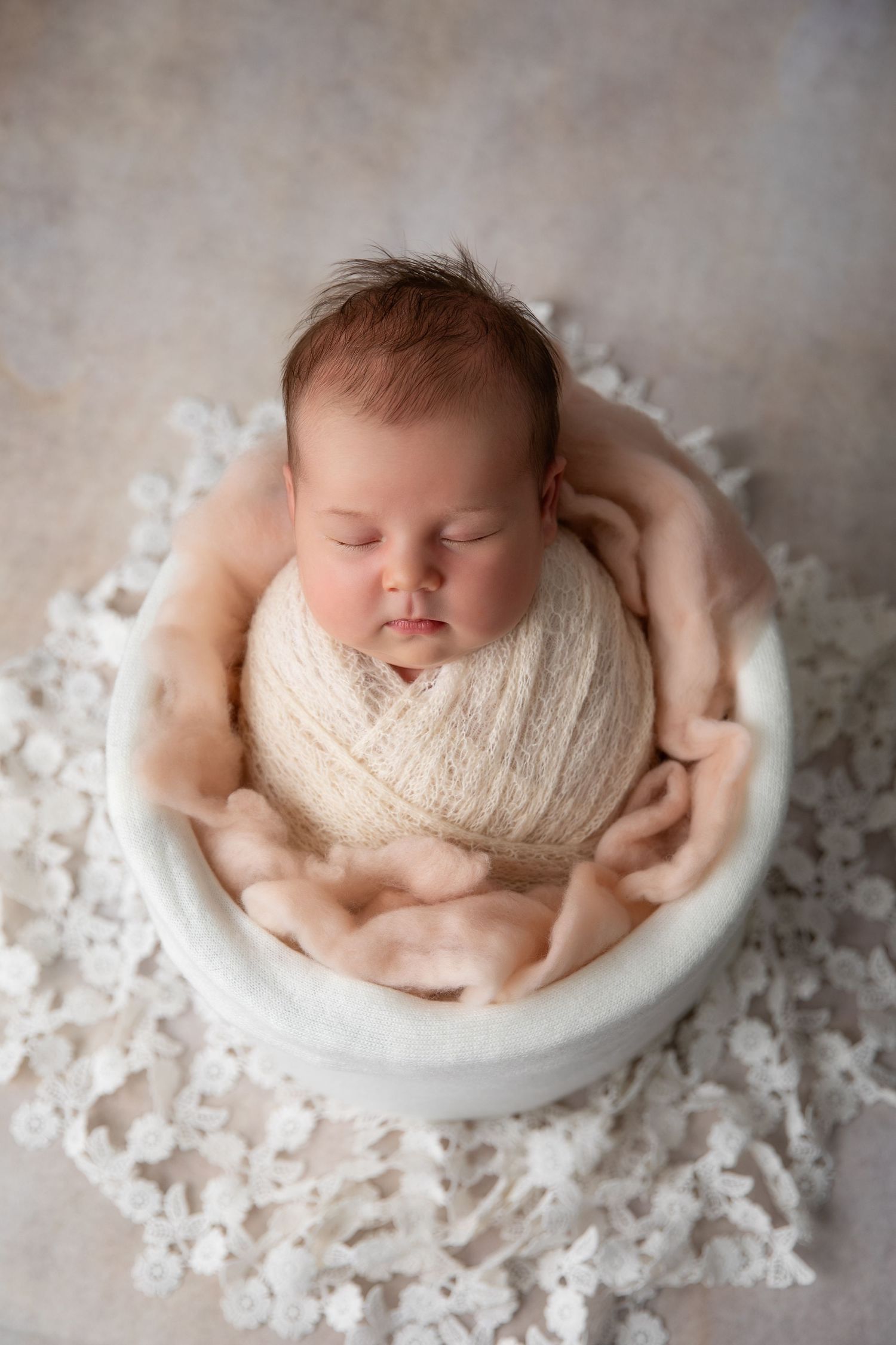 Sleeping newborn wrapped in soft white knit fabric lying in a white bowl surrounded by delicate lace.