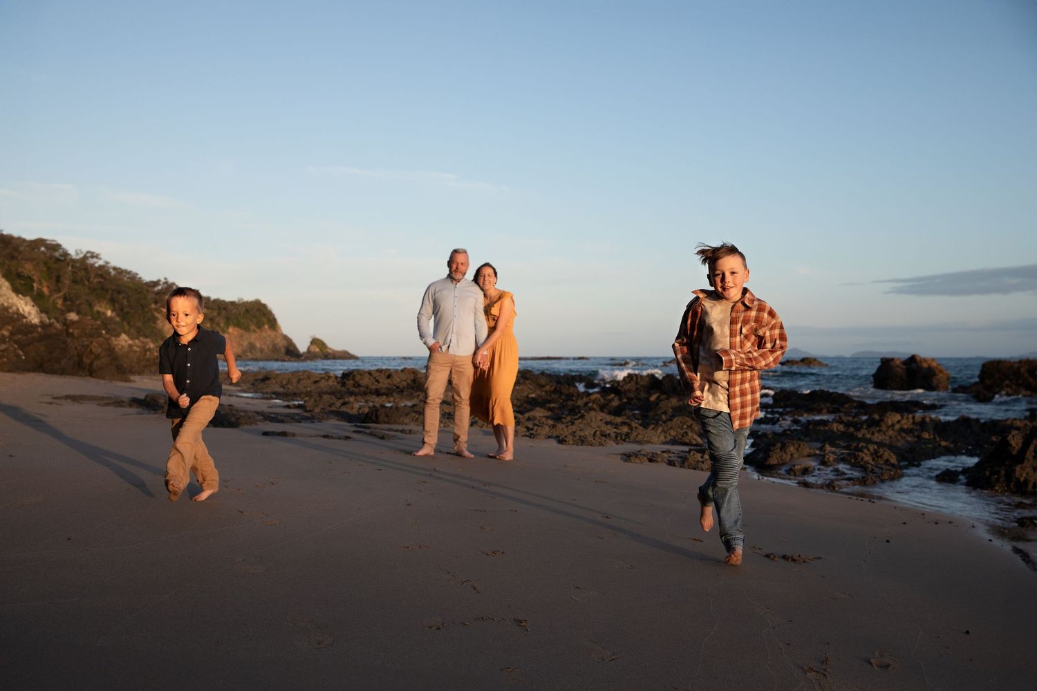 People walking along a scenic beach at sunset with rocky coastline and cliffs in the background.