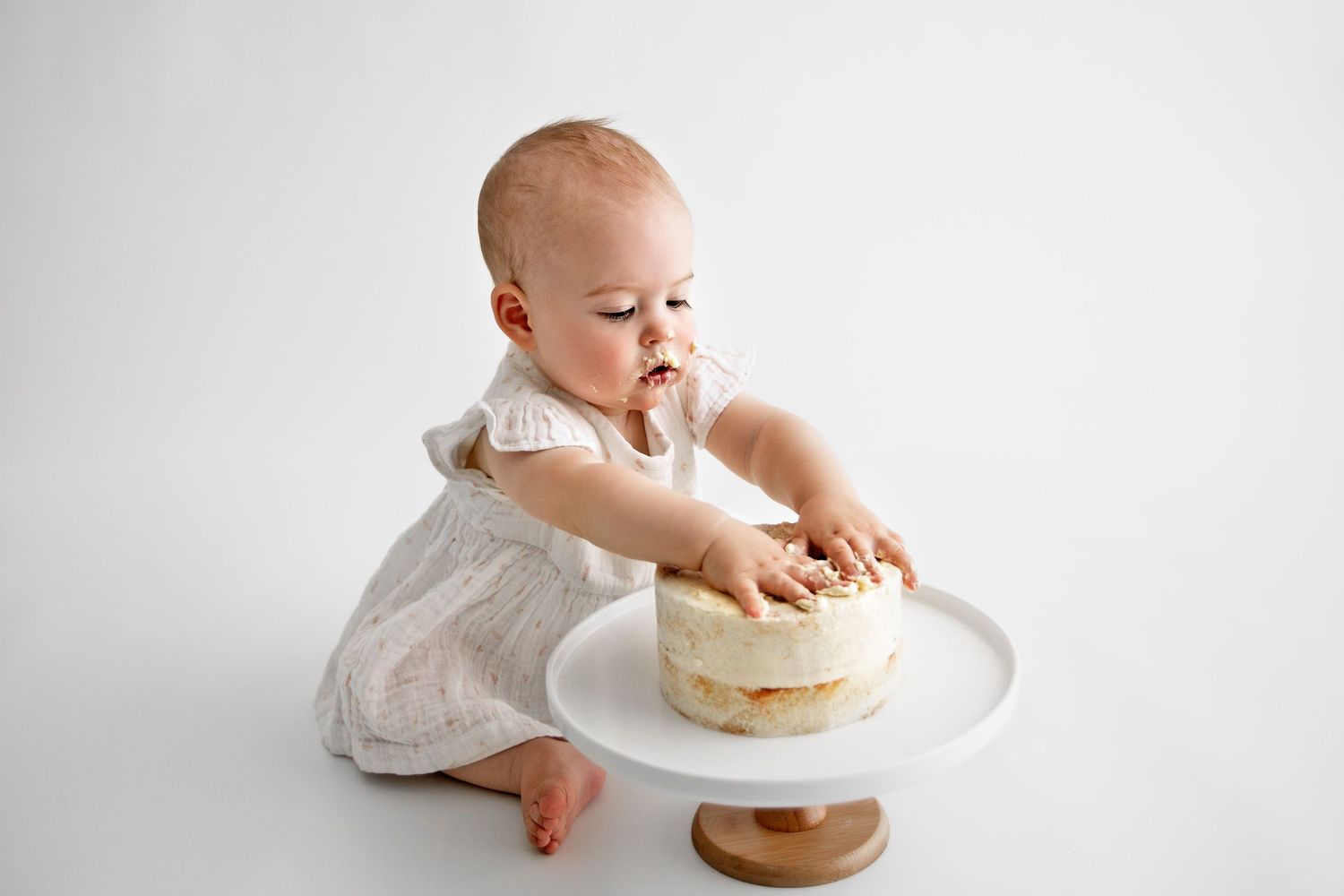 Baby in white dress reaches for cake on plate during photoshoot.