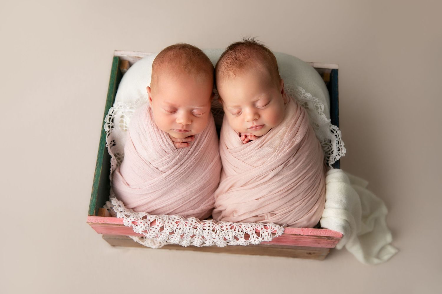 Two sleeping newborns wrapped in soft pink swaddles lie side by side in a cozy white setting.