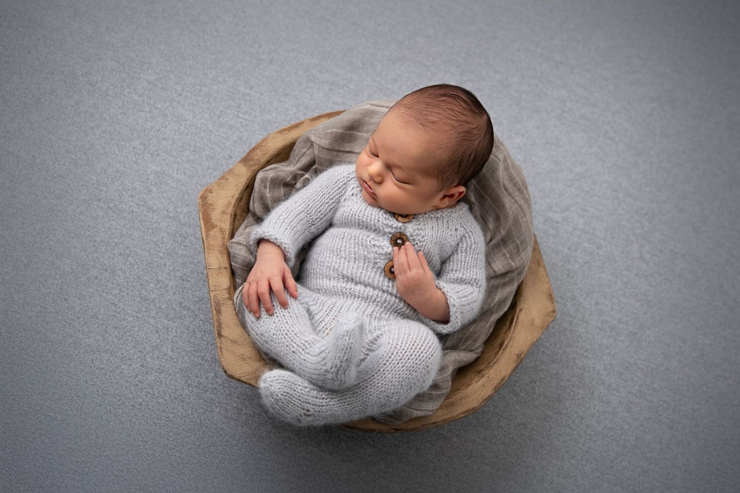 A sleeping newborn baby wearing gray clothing nestles peacefully in a brown woven basket against a gray background.