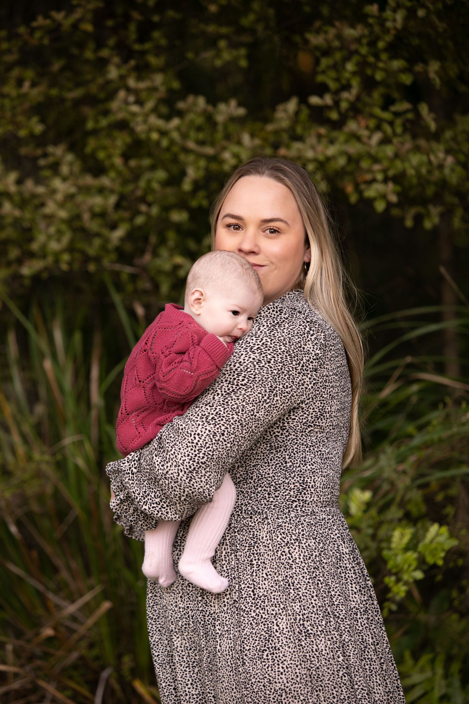A parent in a leopard print dress holds and kisses a baby wearing burgundy in an outdoor garden setting.