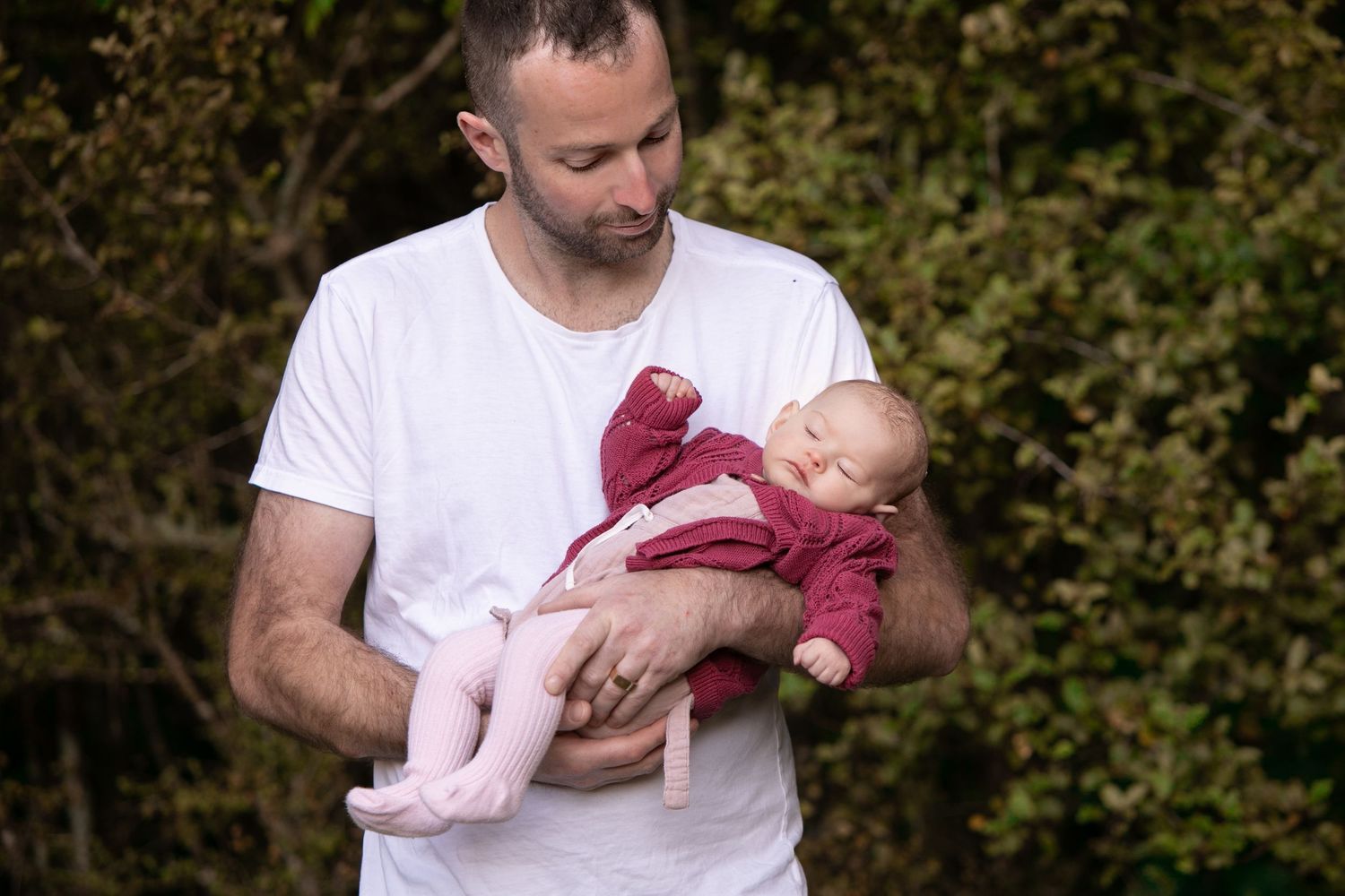 A person in a white t-shirt tenderly holds a sleeping baby wearing pink clothing outdoors against greenery.