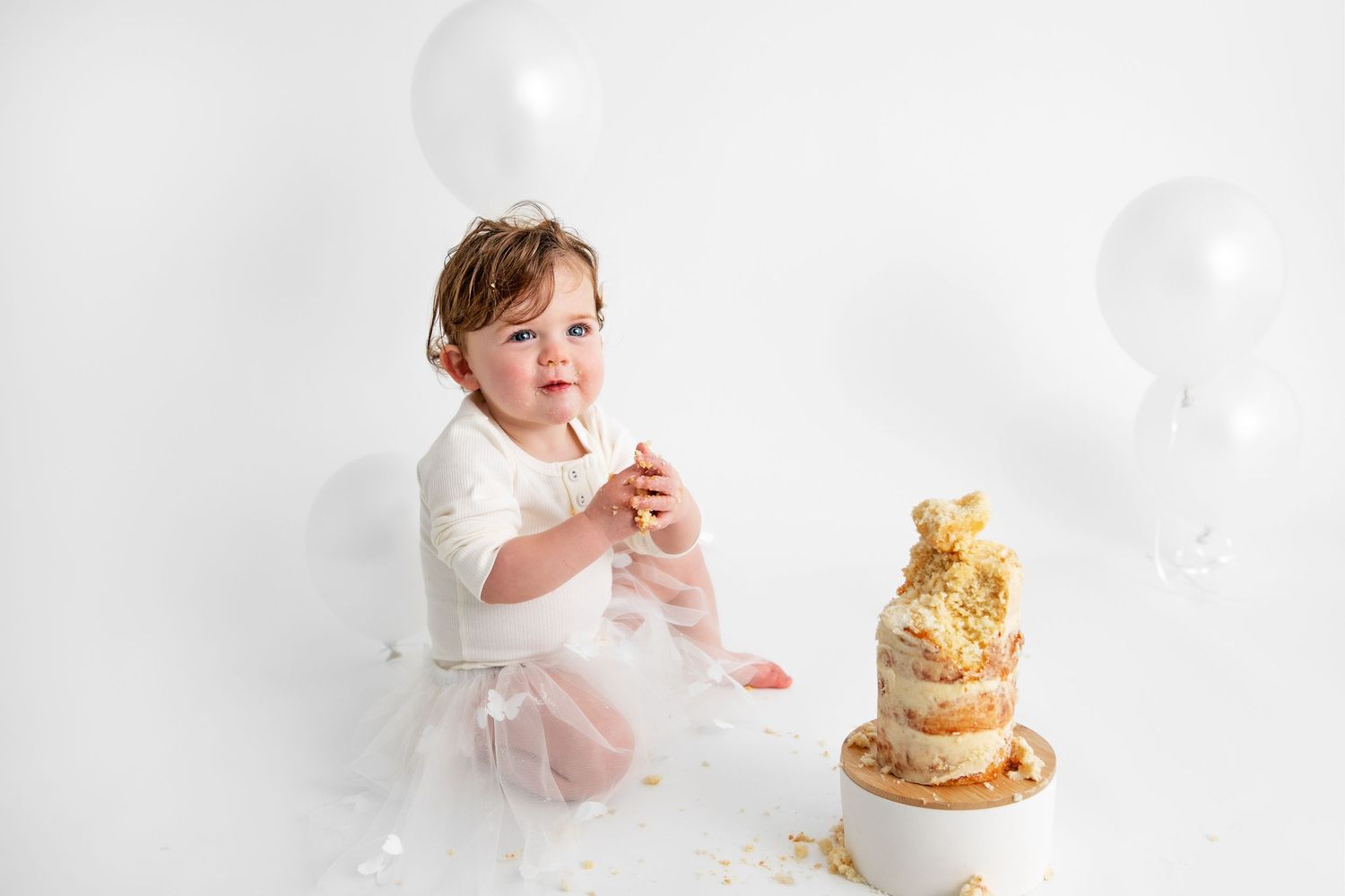 A baby in white clothing sits on the floor with a gold and white birthday cake during a cake smash photo session.