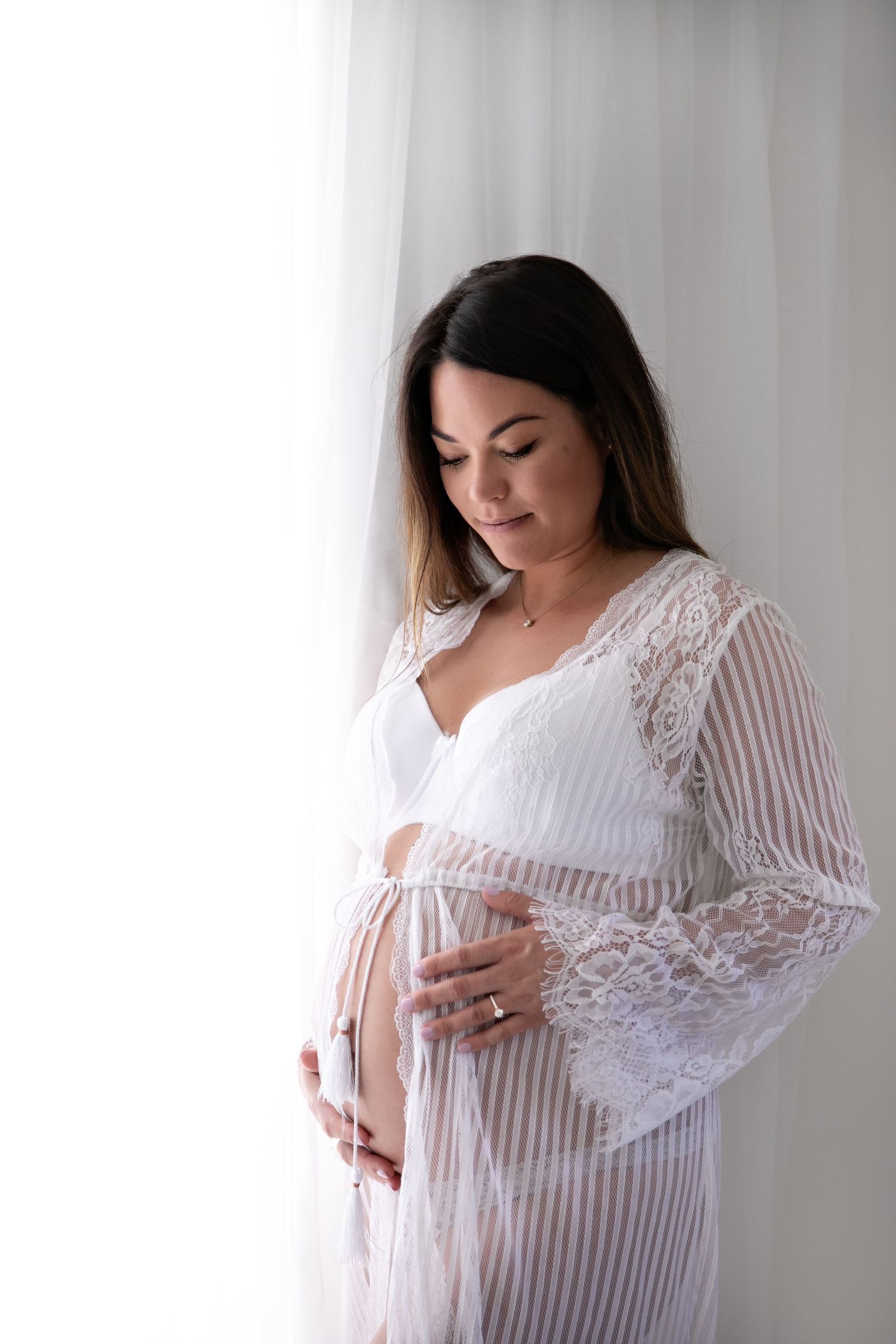 Woman in white lace maternity gown poses against light background in elegant studio portraits.