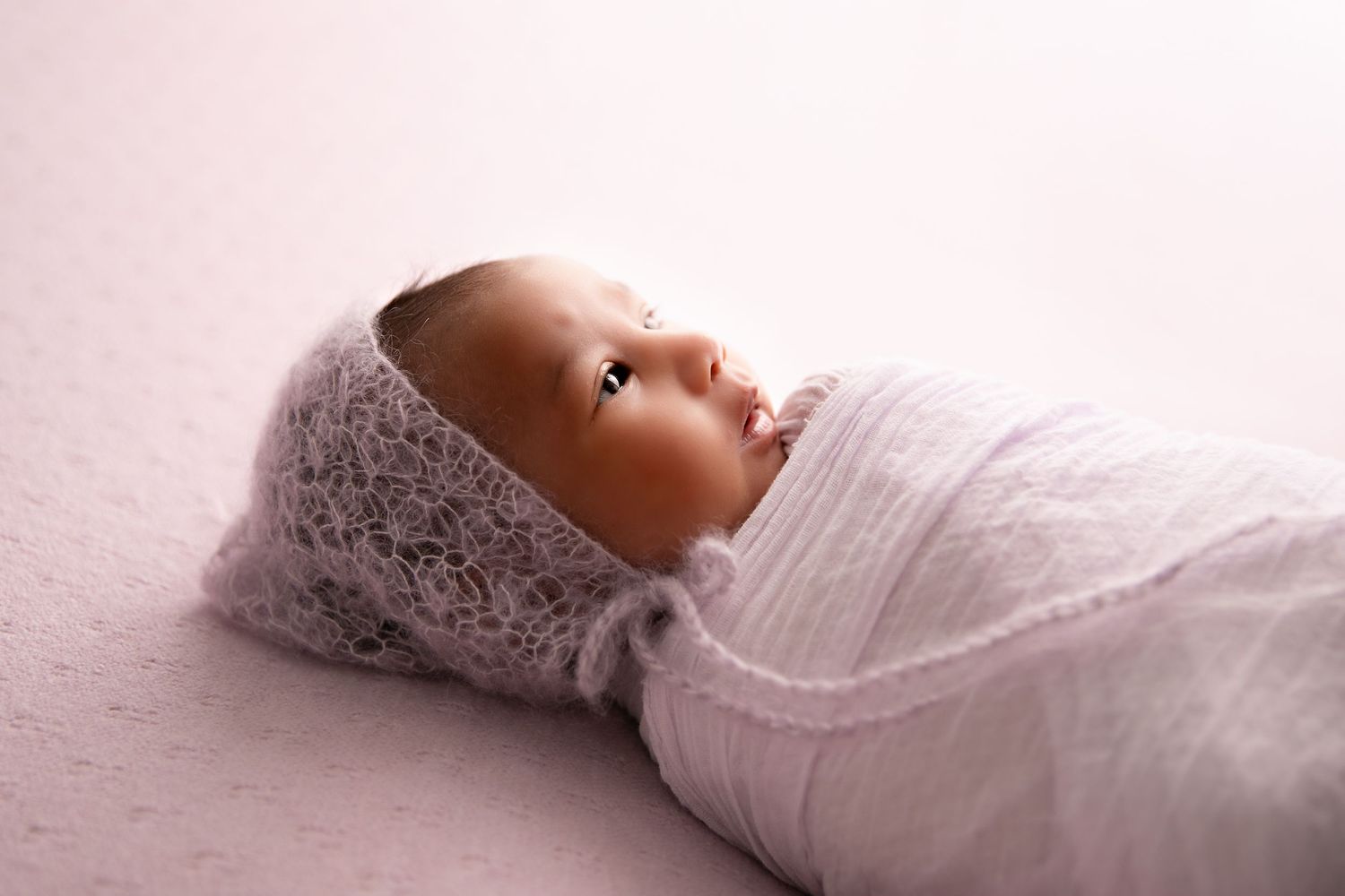 A baby swaddled in white fabric lies on a soft pink surface with delicate lace details.