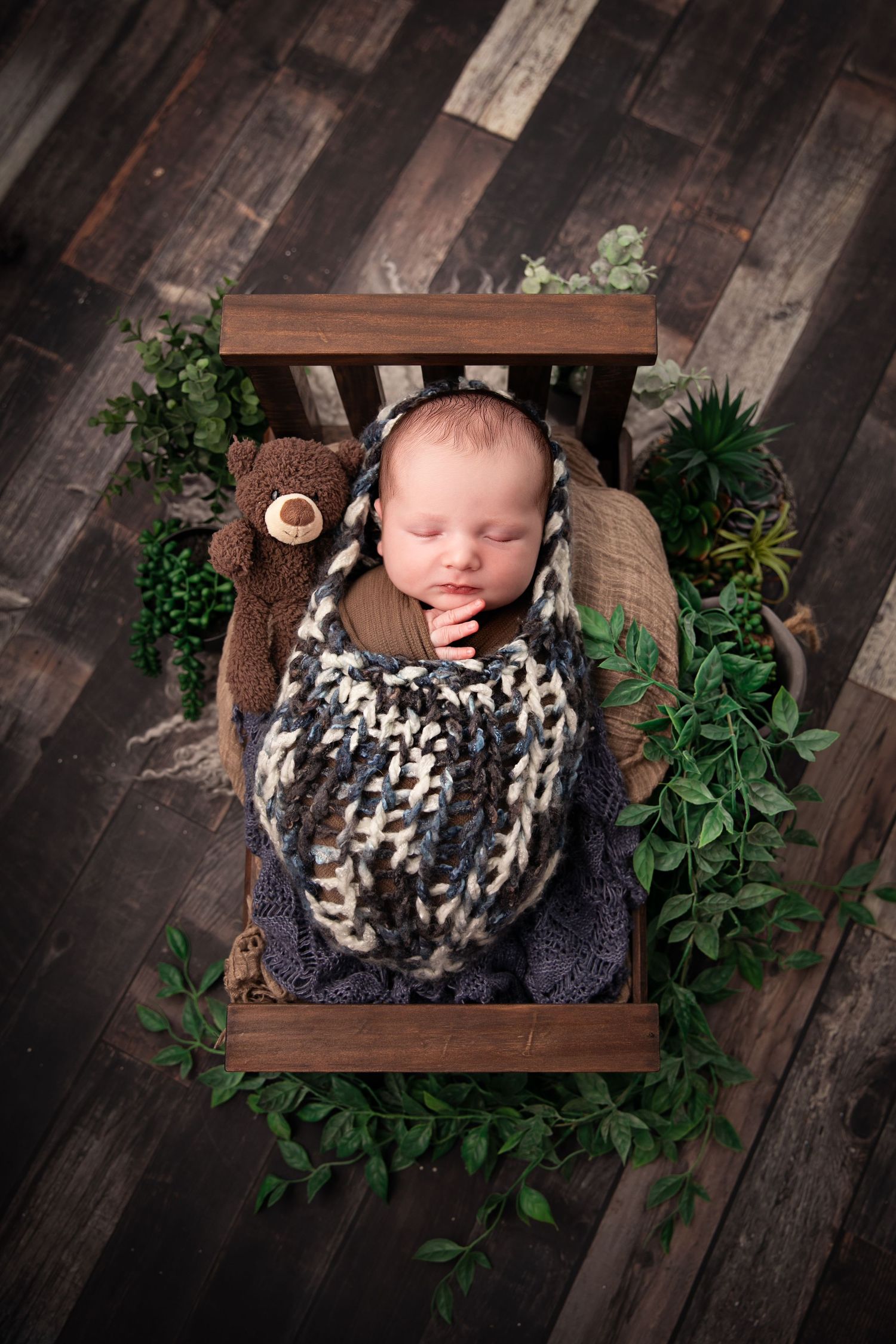 Sleeping newborn in leopard print wrap nestled in wooden crate with greenery on dark wood floor.