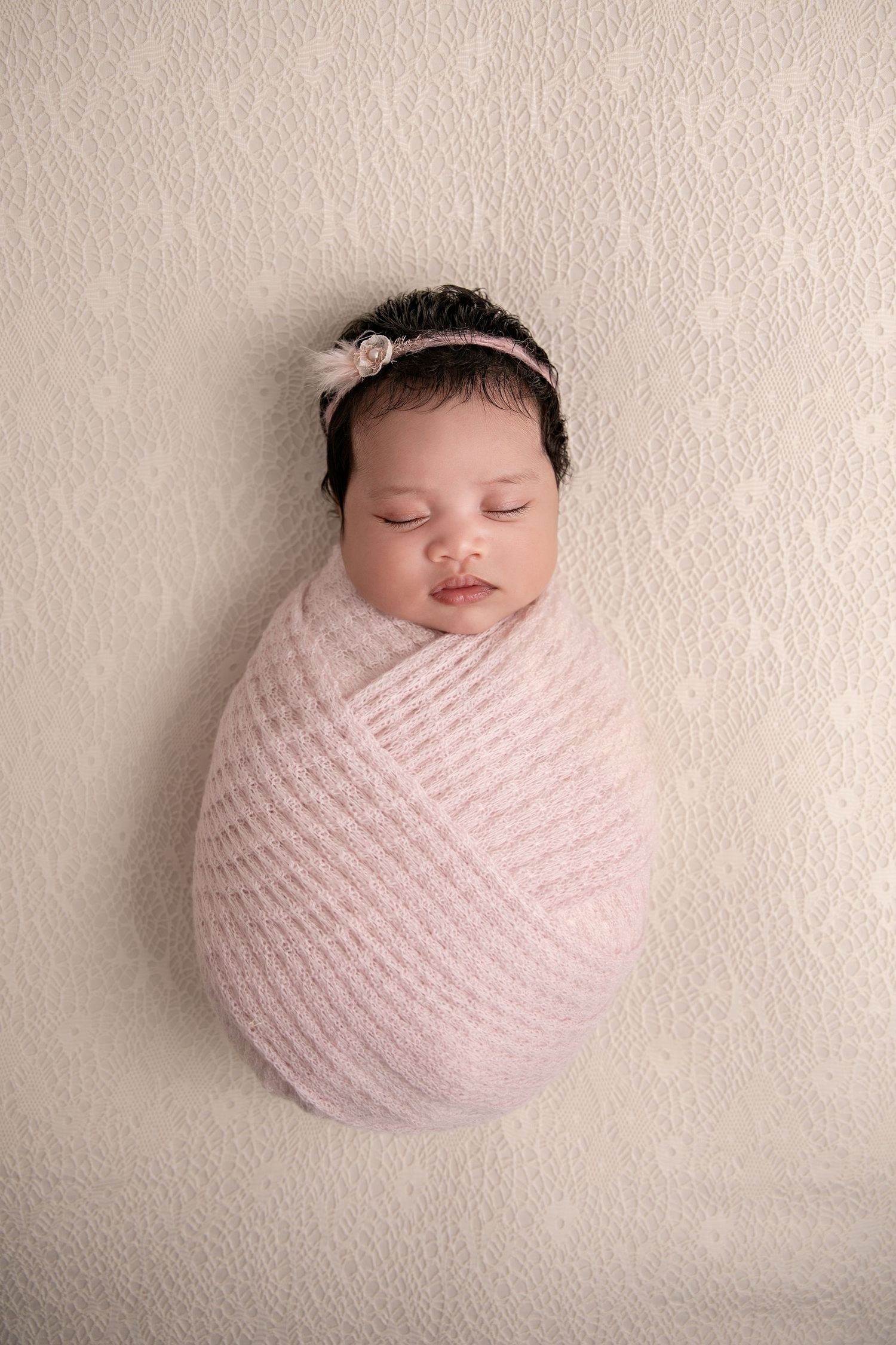 A sleeping newborn baby wrapped in soft pink knit fabric against a cream colored textured backdrop.