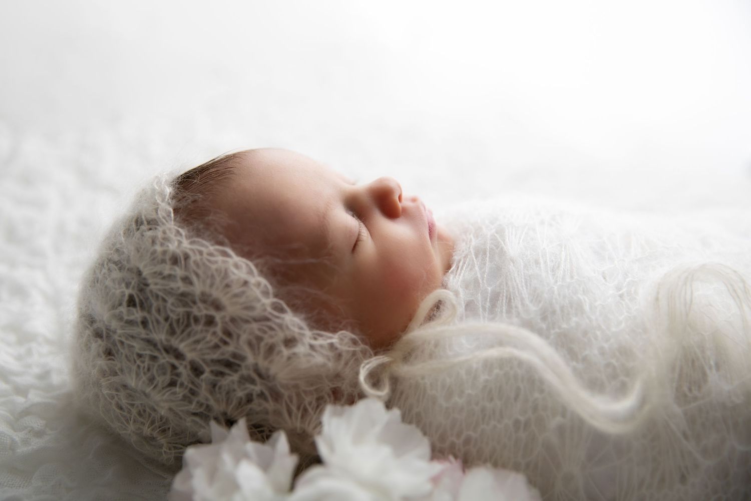 A sleeping baby wrapped in delicate white lace fabric lies peacefully against a light background.