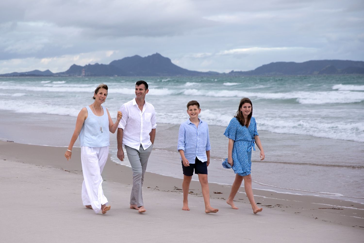 Family walking together on beach at sunset holding hands with ocean waves and mountains in background.