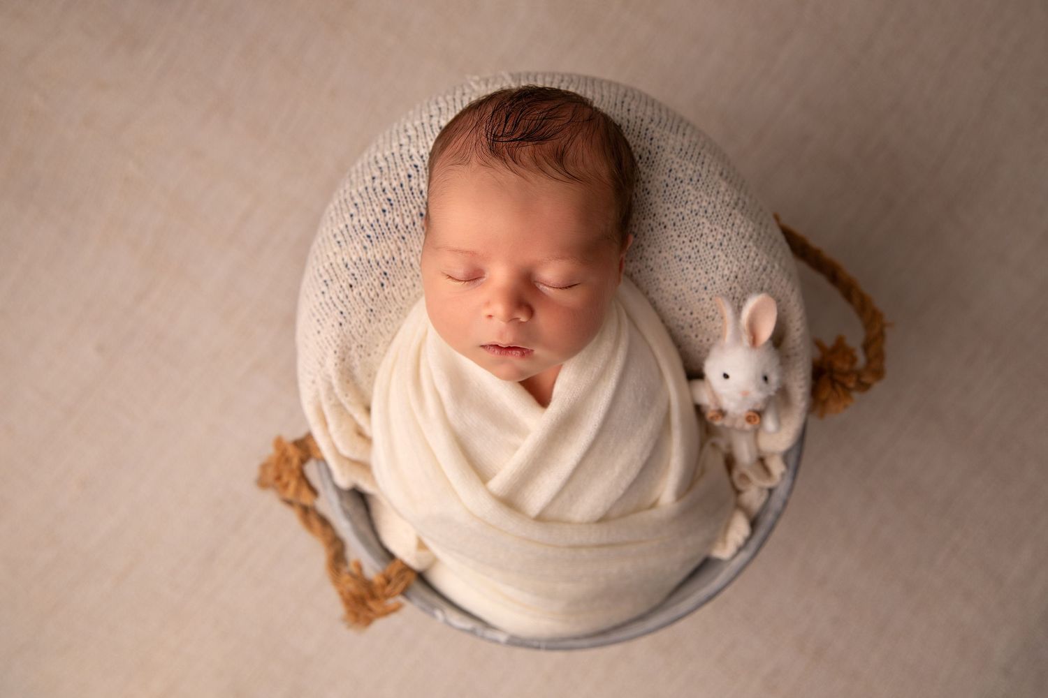 Sleeping newborn baby wrapped in white fabric lies on beige background with soft stuffed bunny toy.