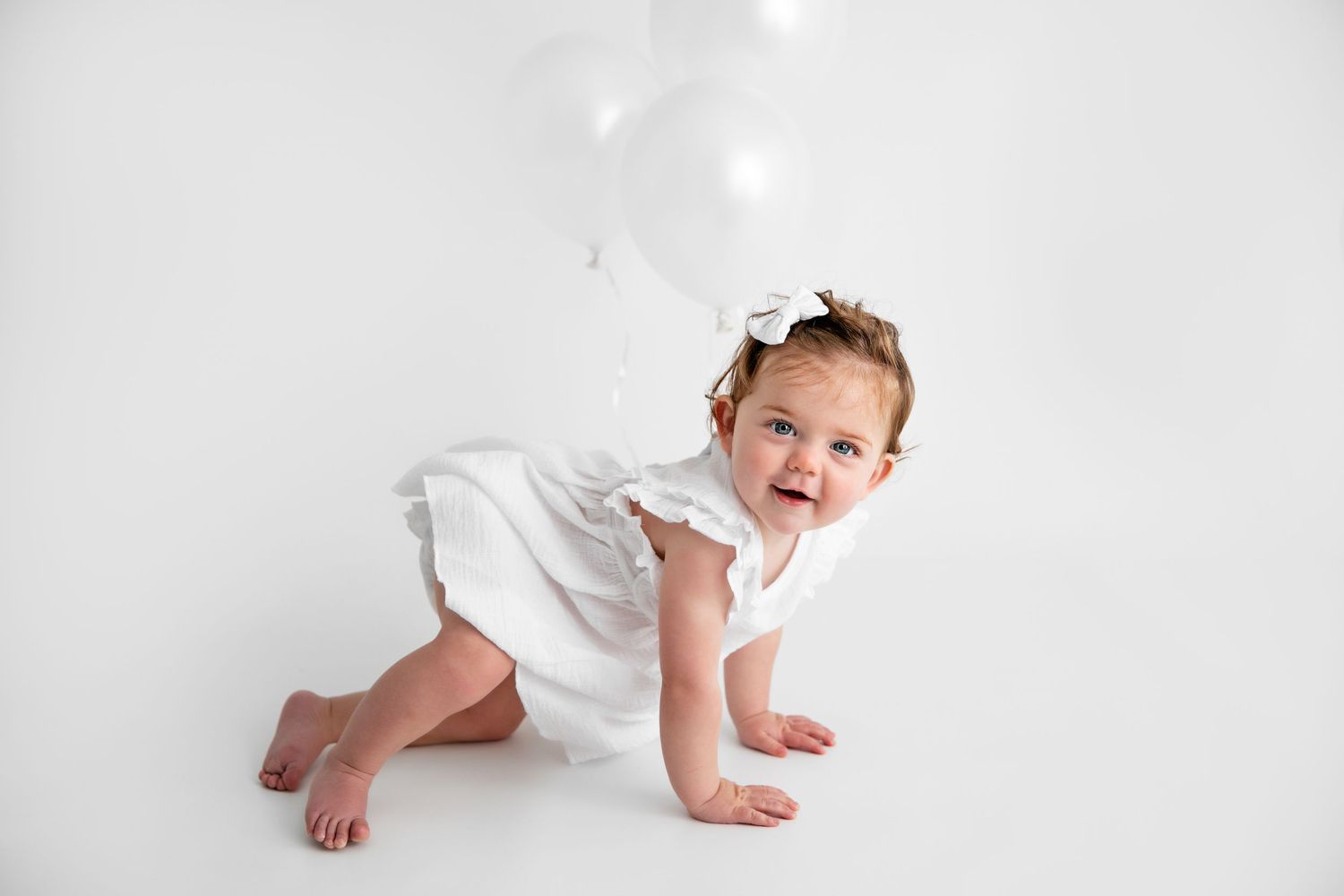 Baby in white outfit crawls and smiles on white background during studio photo session.