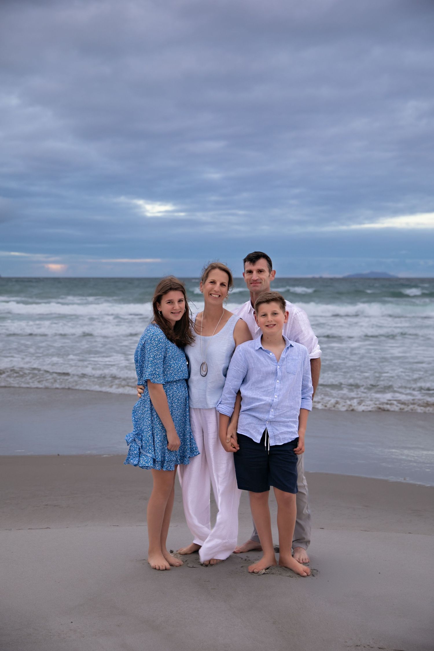 Family group poses together on cloudy beach with waves and mountains in background.