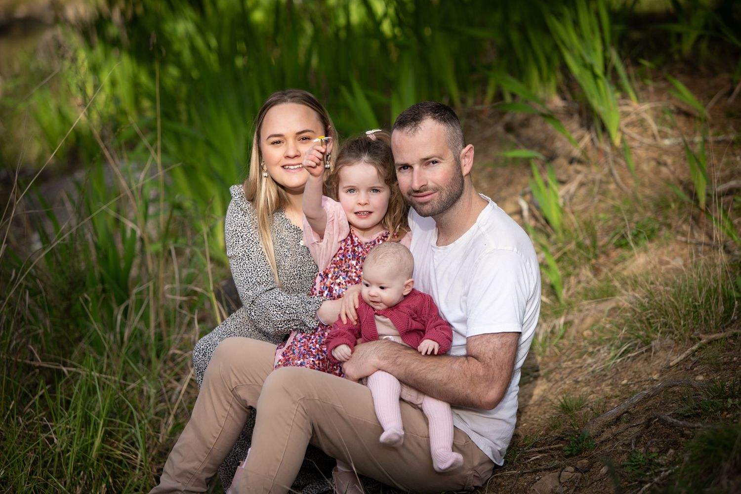 A family wearing casual clothing sits together on a grassy hillside during an outdoor photo session.