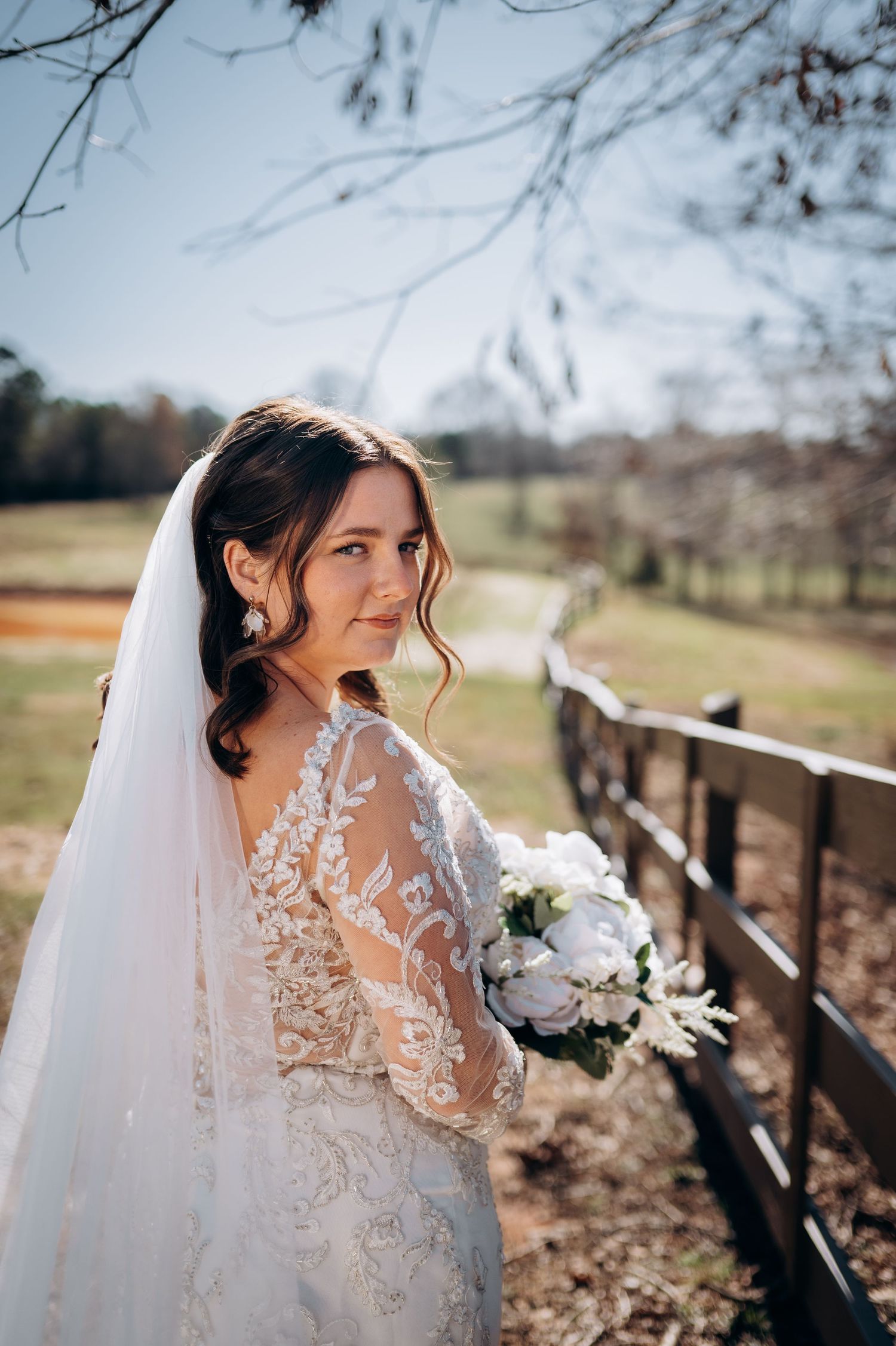 Bride in lace long-sleeve wedding gown and veil holds white bouquet by rustic fence in sunny outdoor setting.