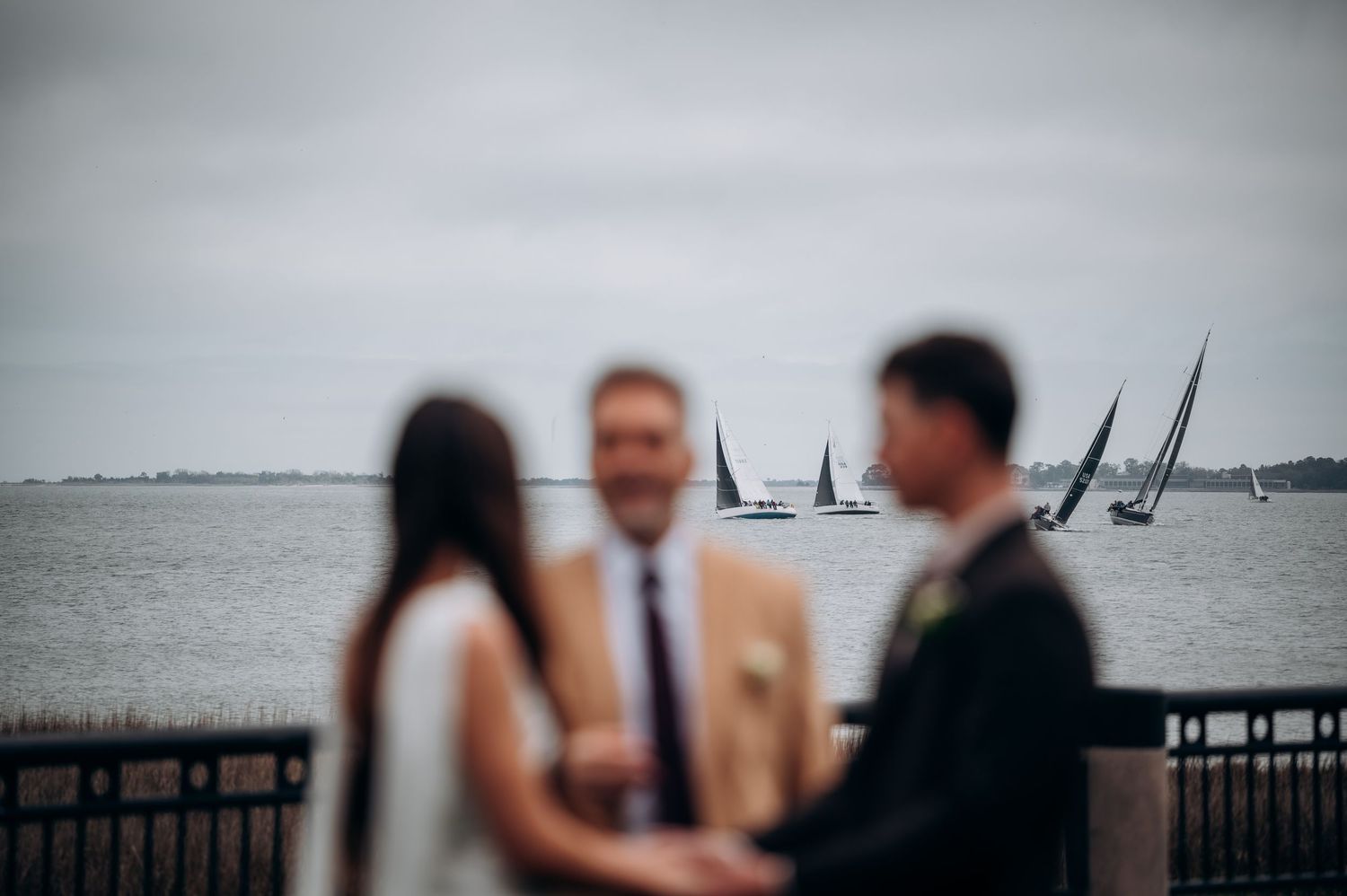 Blurred couple in foreground with officiant during outdoor waterfront wedding ceremony, sailboats visible on the bay.