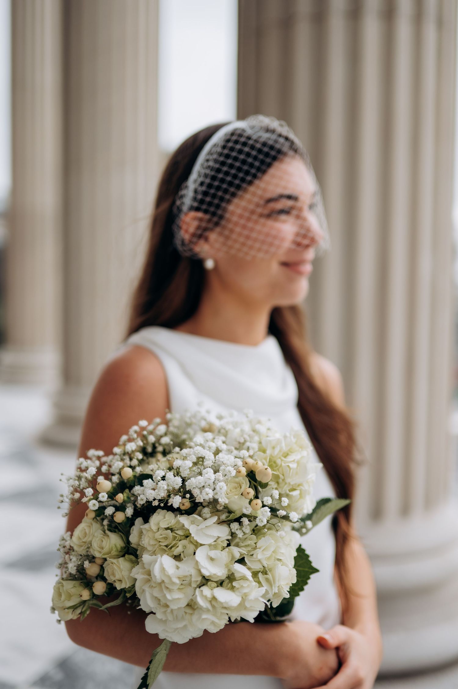 Elegant bride in white dress wearing birdcage veil holding white hydrangea and baby's breath bouquet by columns.