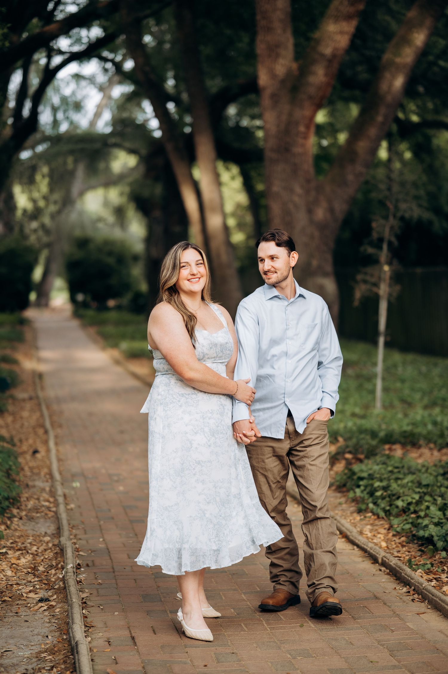 Couple holding hands and smiling while walking on a brick path surrounded by lush green trees.