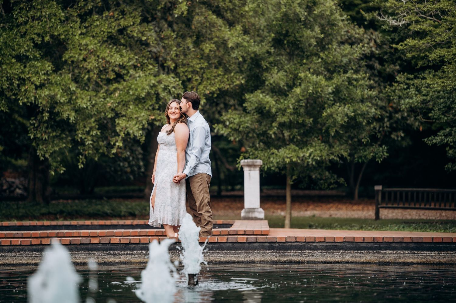 Romantic couple standing close together by a garden fountain with lush green trees in the background.