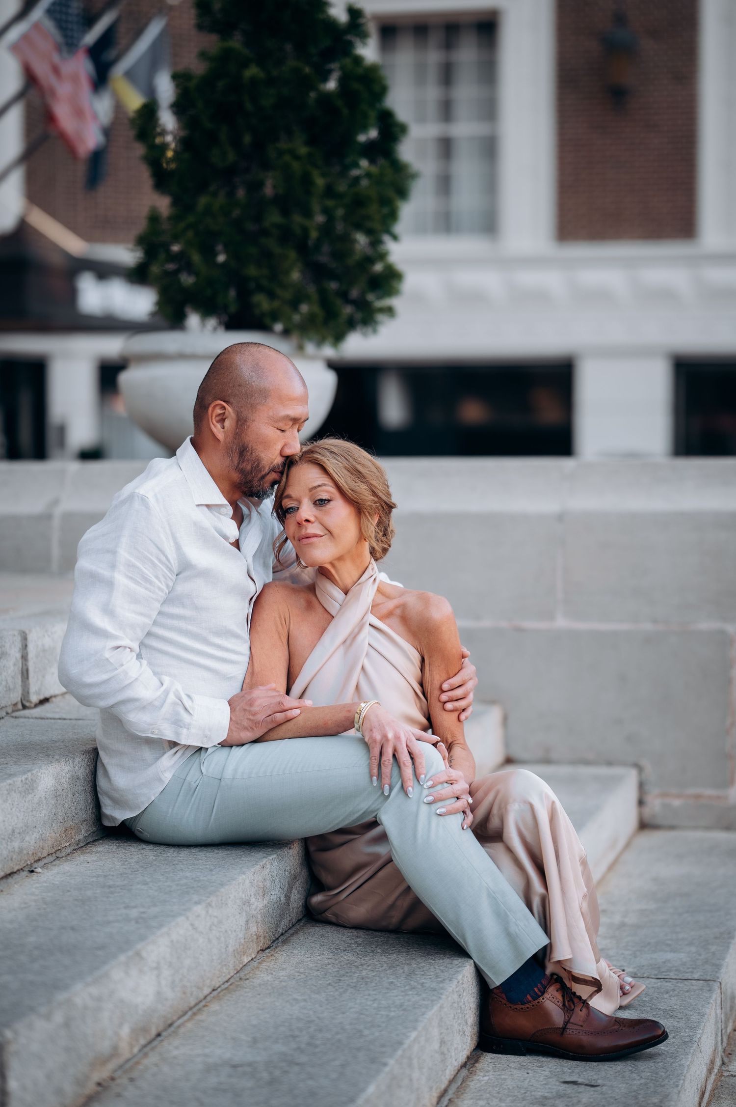 Couple sitting closely on outdoor stone steps, man in white shirt embracing woman in blush outfit.