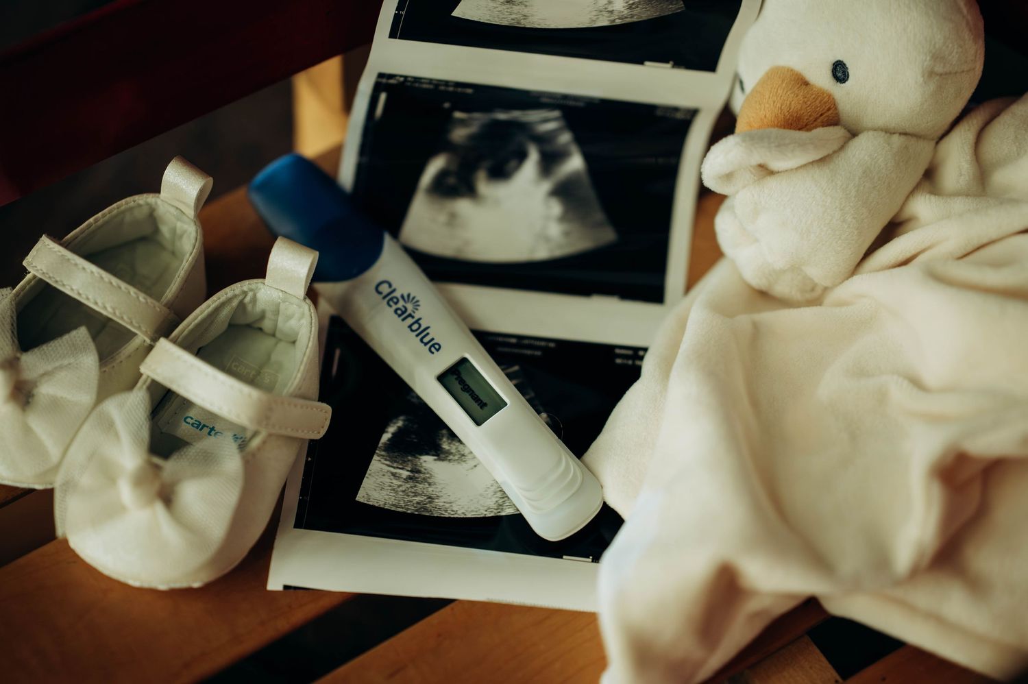 Baby shoes, ultrasound photos, pregnancy test, and duck plush toy arranged on wooden surface announcing a new pregnancy.