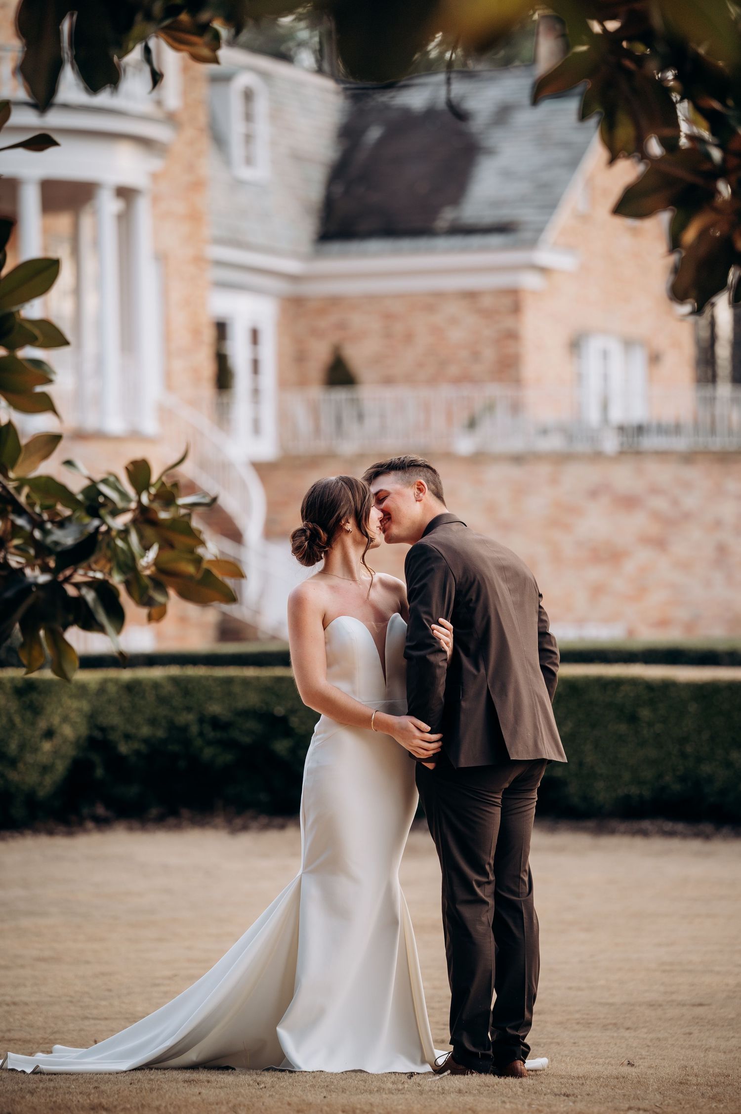 Newlyweds share a kiss in front of elegant stone mansion surrounded by manicured gardens.
