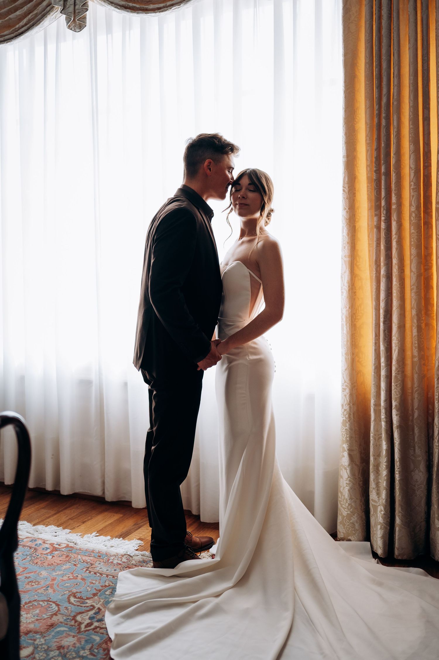 Bride and groom sharing intimate moment in silhouette before large window with flowing white curtains.