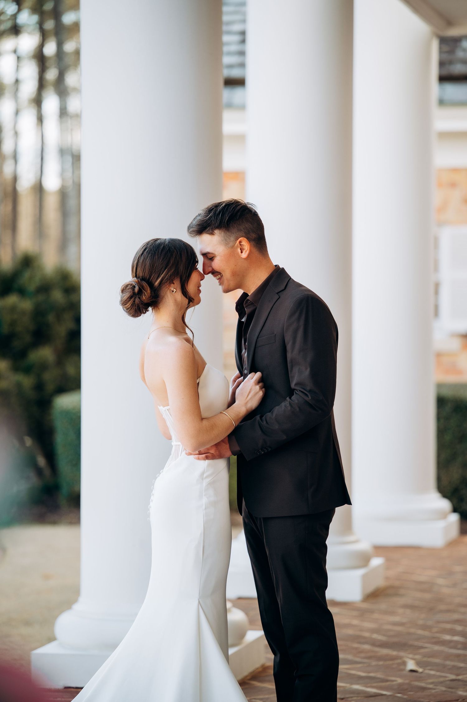 Bride and groom share an intimate moment embracing between white columns outdoors.