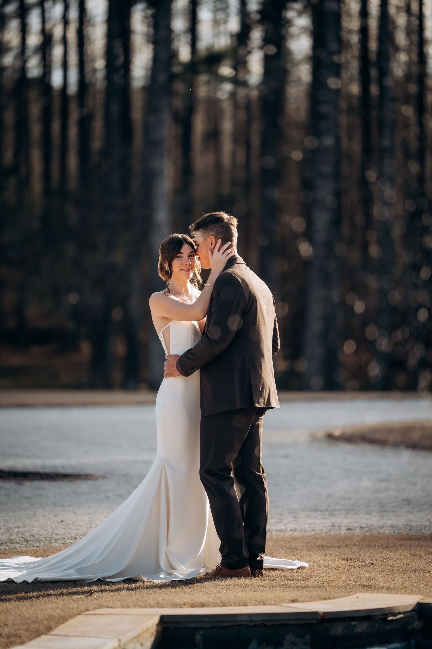 Bride and groom embracing tenderly outdoors with a sunlit forest backdrop.