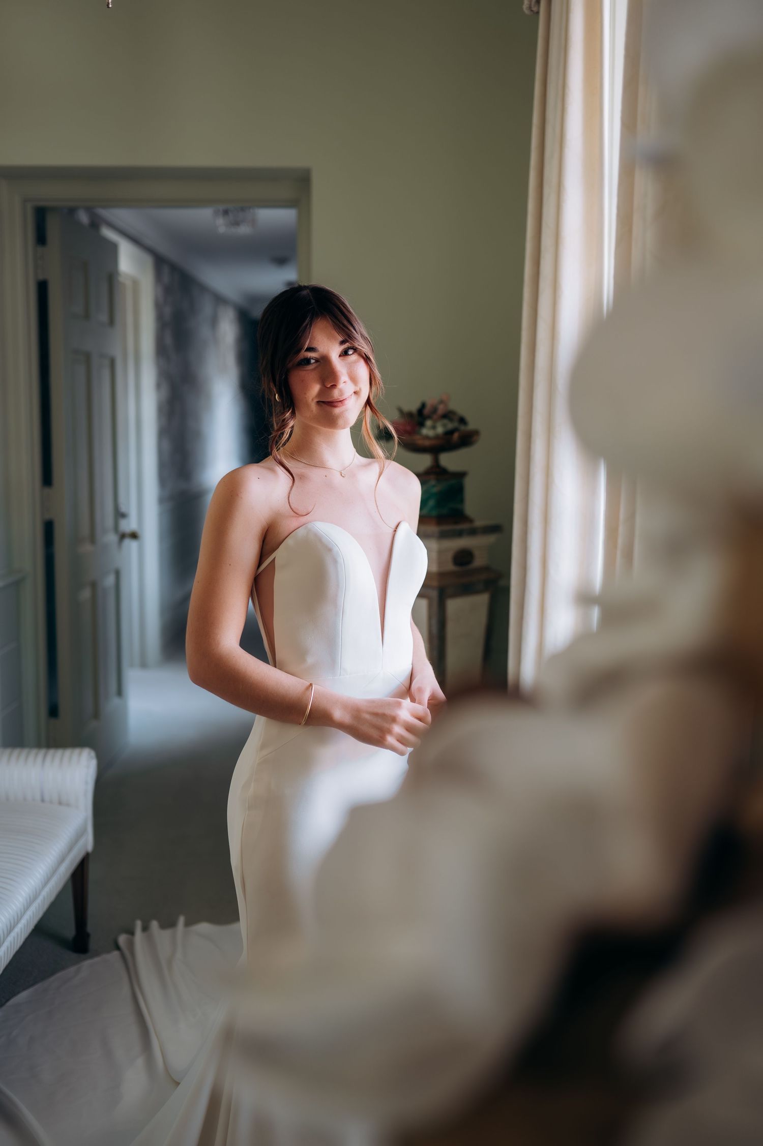 Bride in elegant white sleeveless wedding gown smiling softly near a bright window indoors.