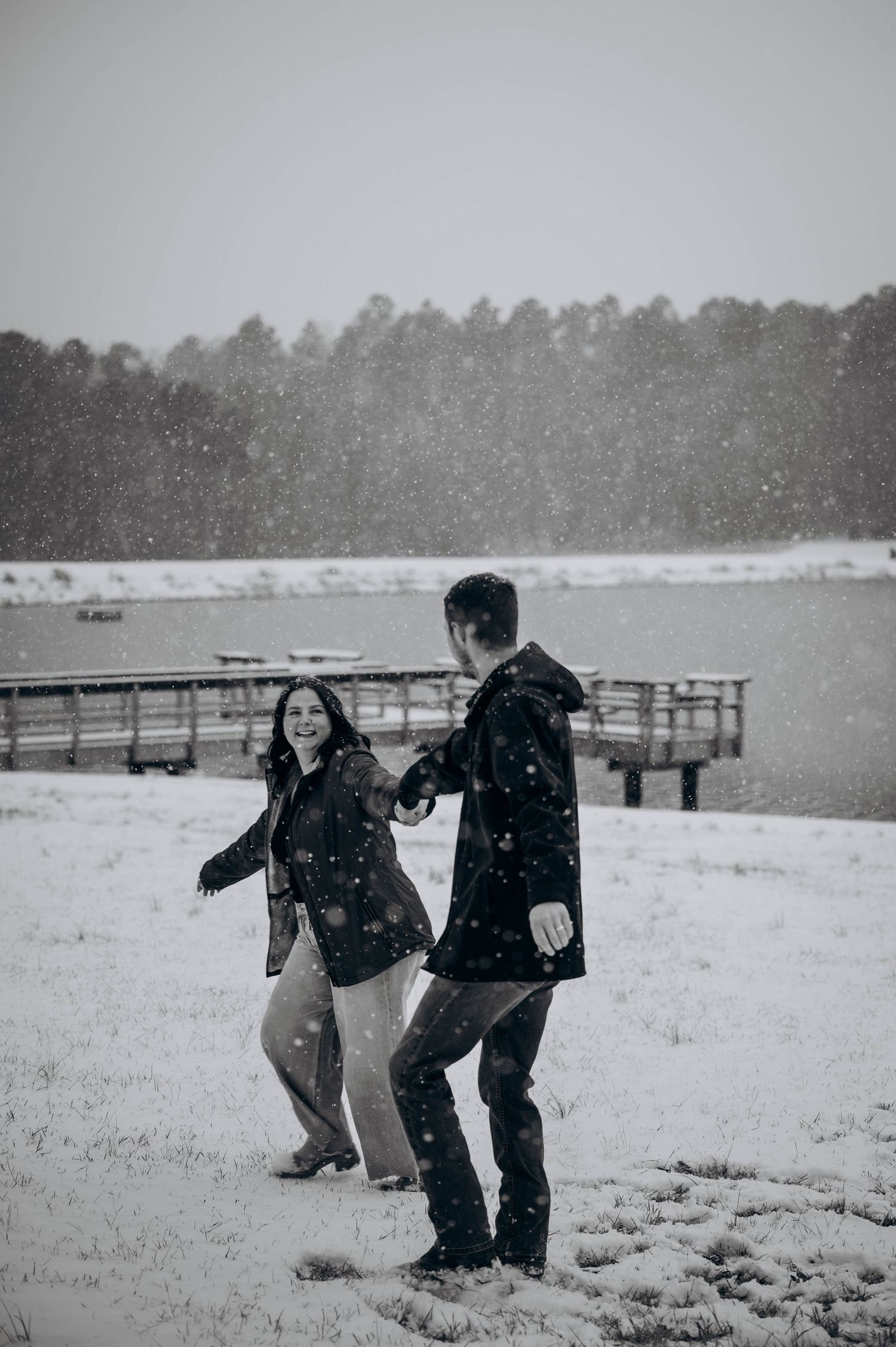 A playful couple enjoys a snowy day together in a winter field with a wooden fence in the background.