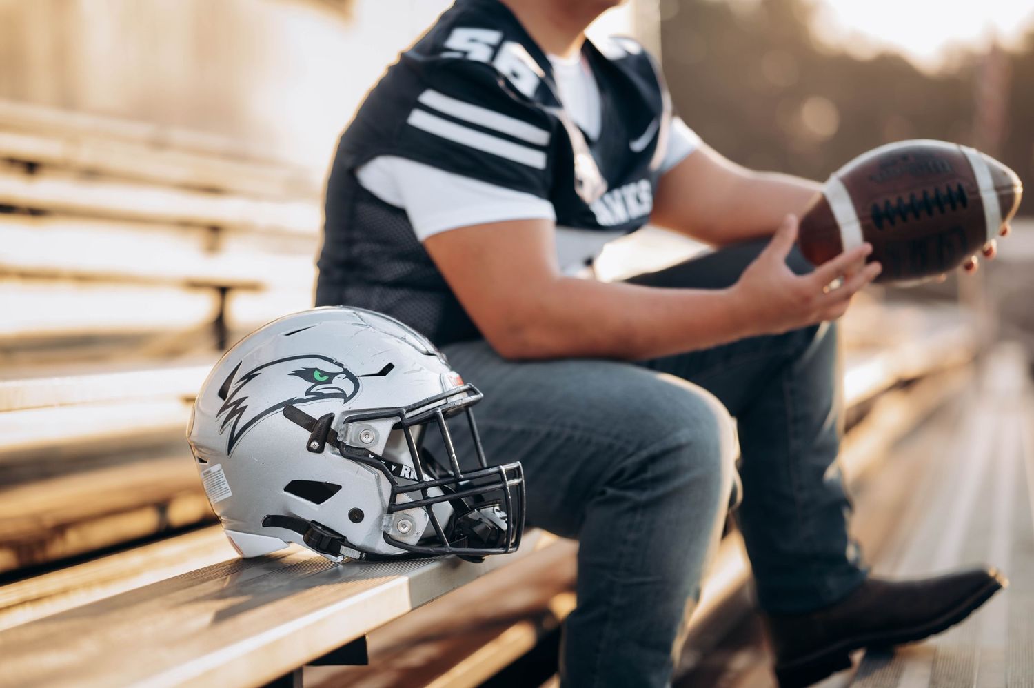 Football player in uniform sitting on stadium bleachers with helmet and ball.
