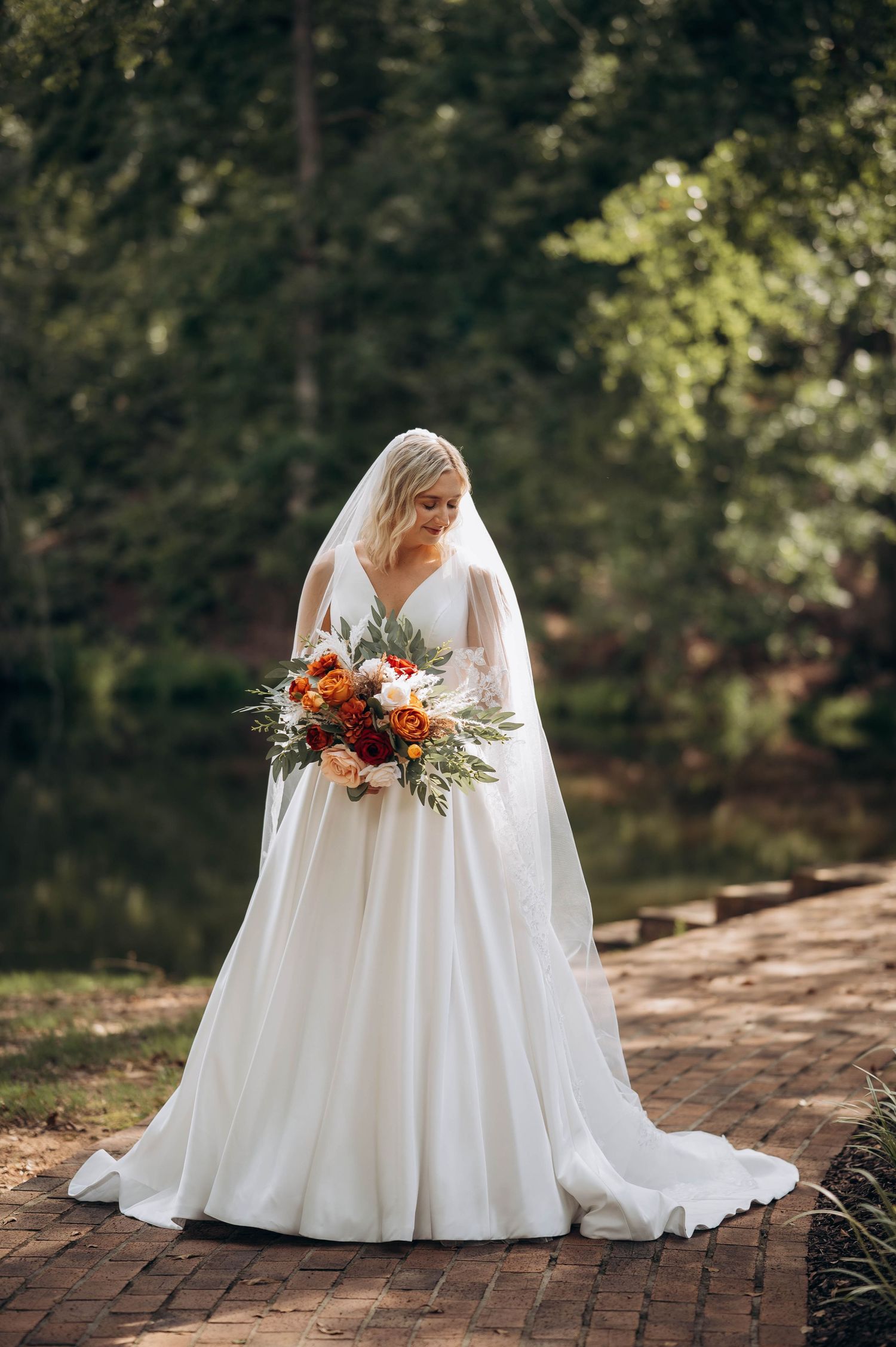 Bride in elegant white dress poses with orange and white bouquet on brick pathway.