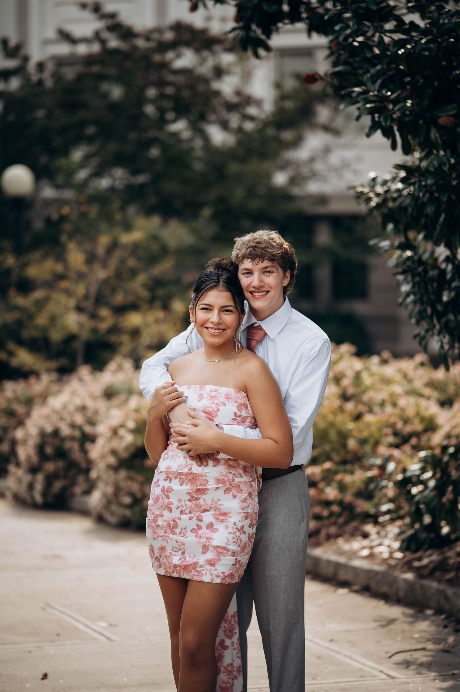 A couple embraces on a garden path with the person wearing a pink floral dress and white shirt against greenery.