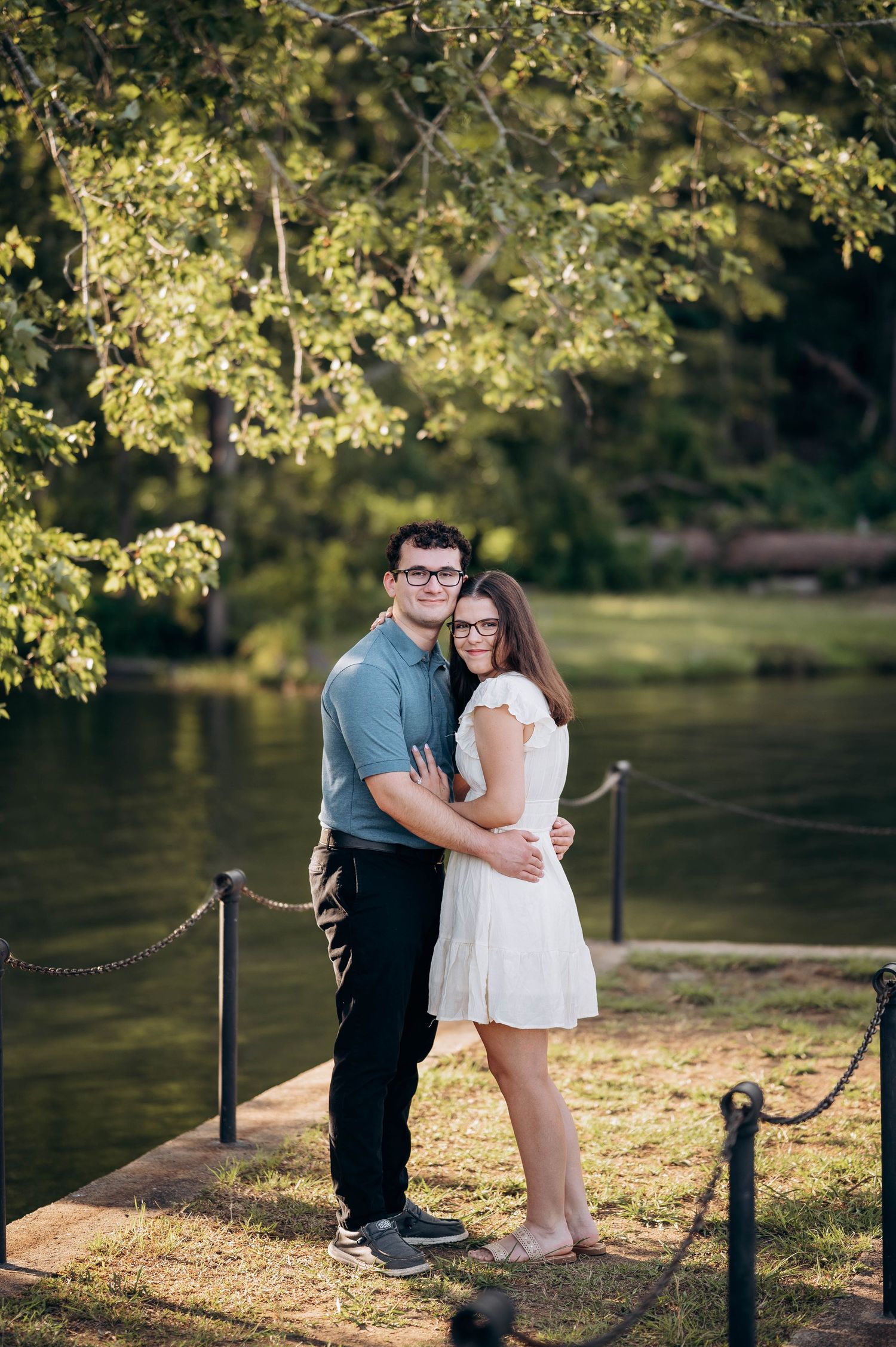 Sweet couple shares tender moment by waterfront with overhanging trees.
