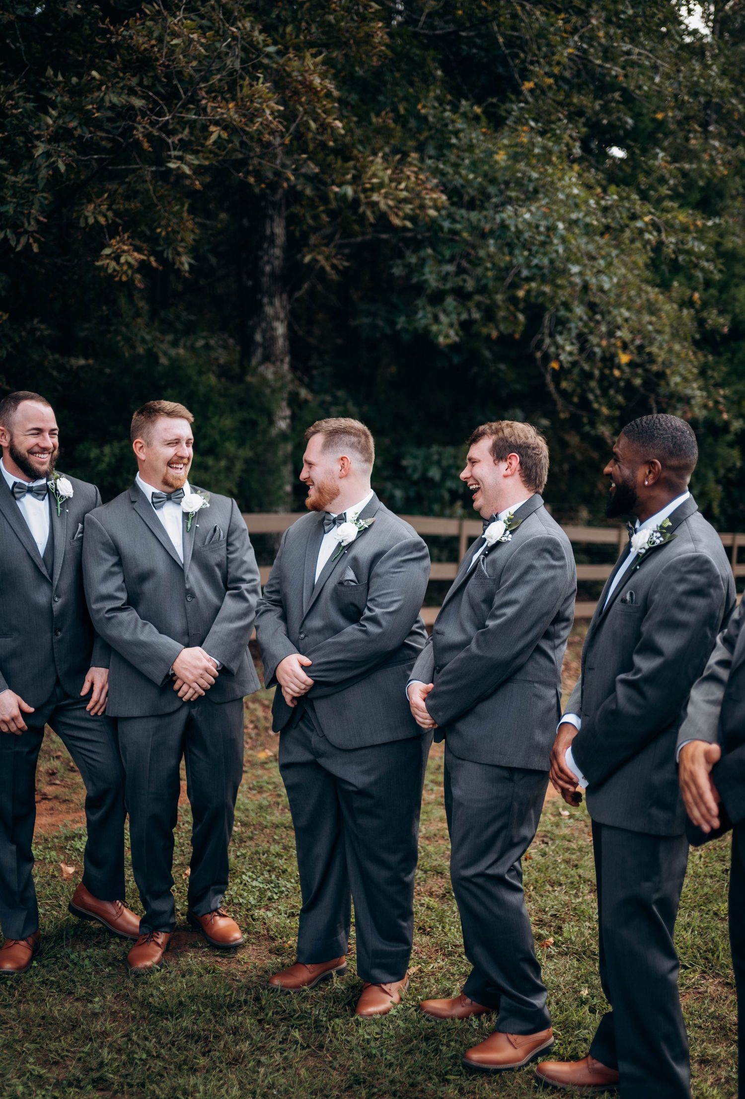 Groomsmen in dark suits show off their brown leather shoes while standing in a line outdoors.