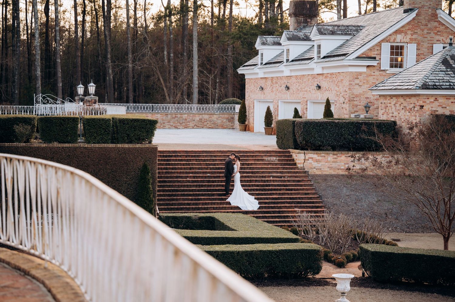 Bride in white gown poses on grand brick staircase surrounded by manicured hedges at elegant estate venue.