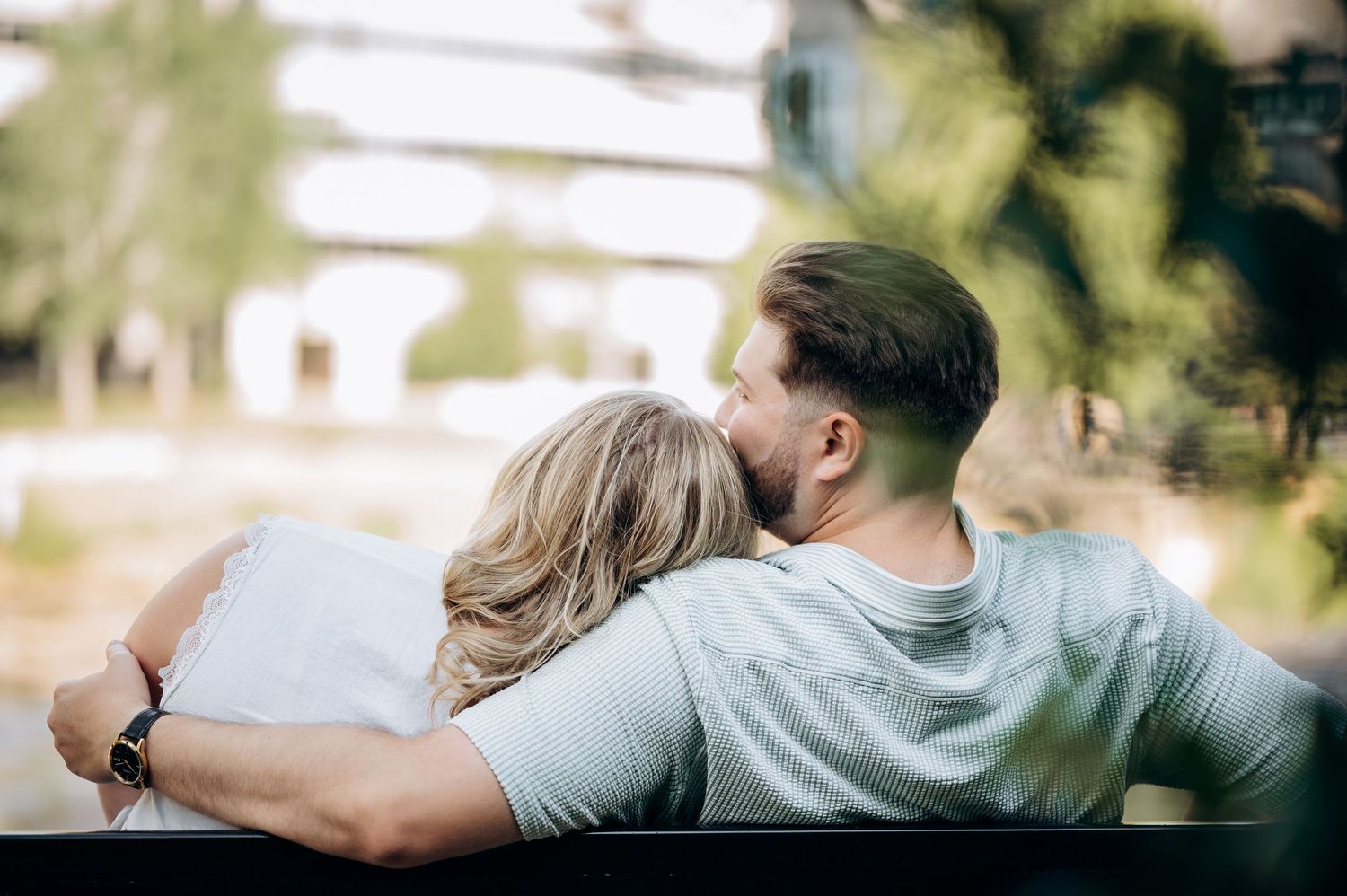 Couple sharing a tender moment on a bench outdoors, man embracing woman from behind in a romantic setting.