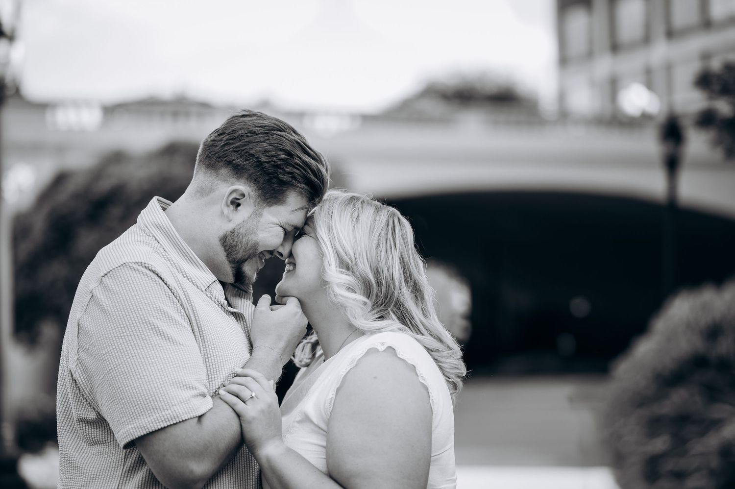 Black and white photo of a romantic couple sharing an intimate moment near a bridge outdoors.