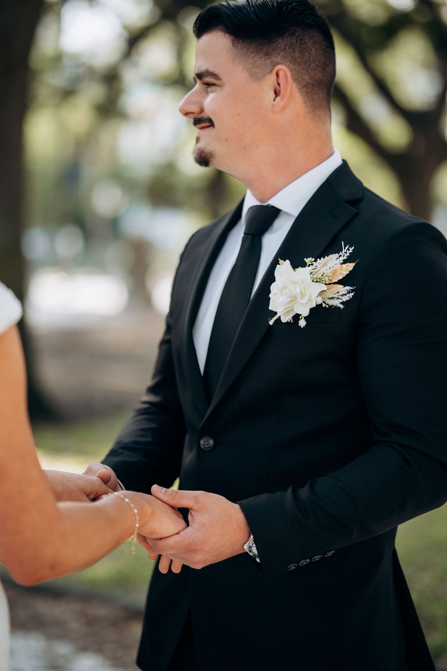 Groom in black tuxedo holds bride's hands during outdoor wedding ceremony with floral boutonniere.