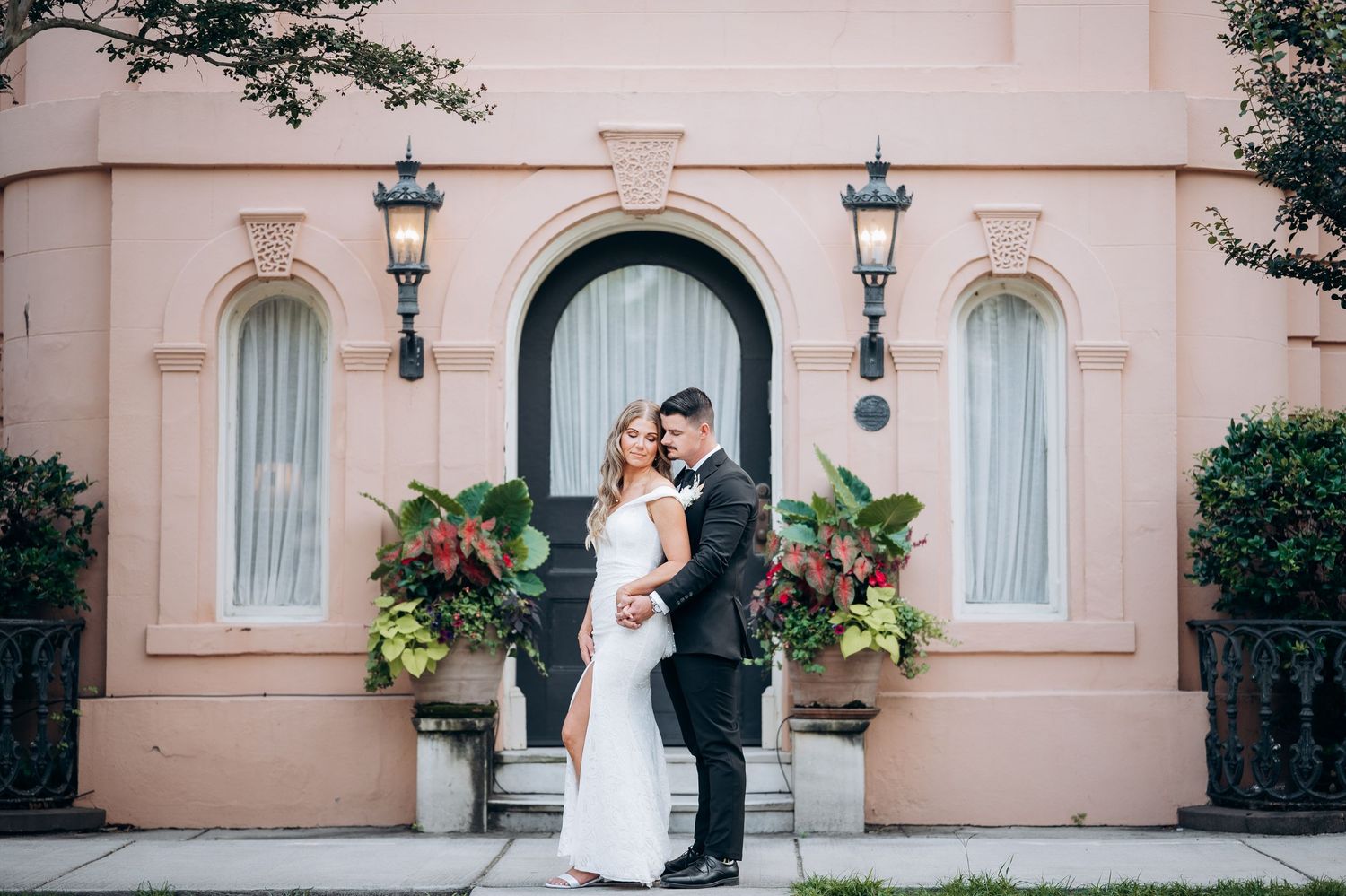 Bride and groom embracing in front of a elegant pink building with arched doorway and lush floral planters.