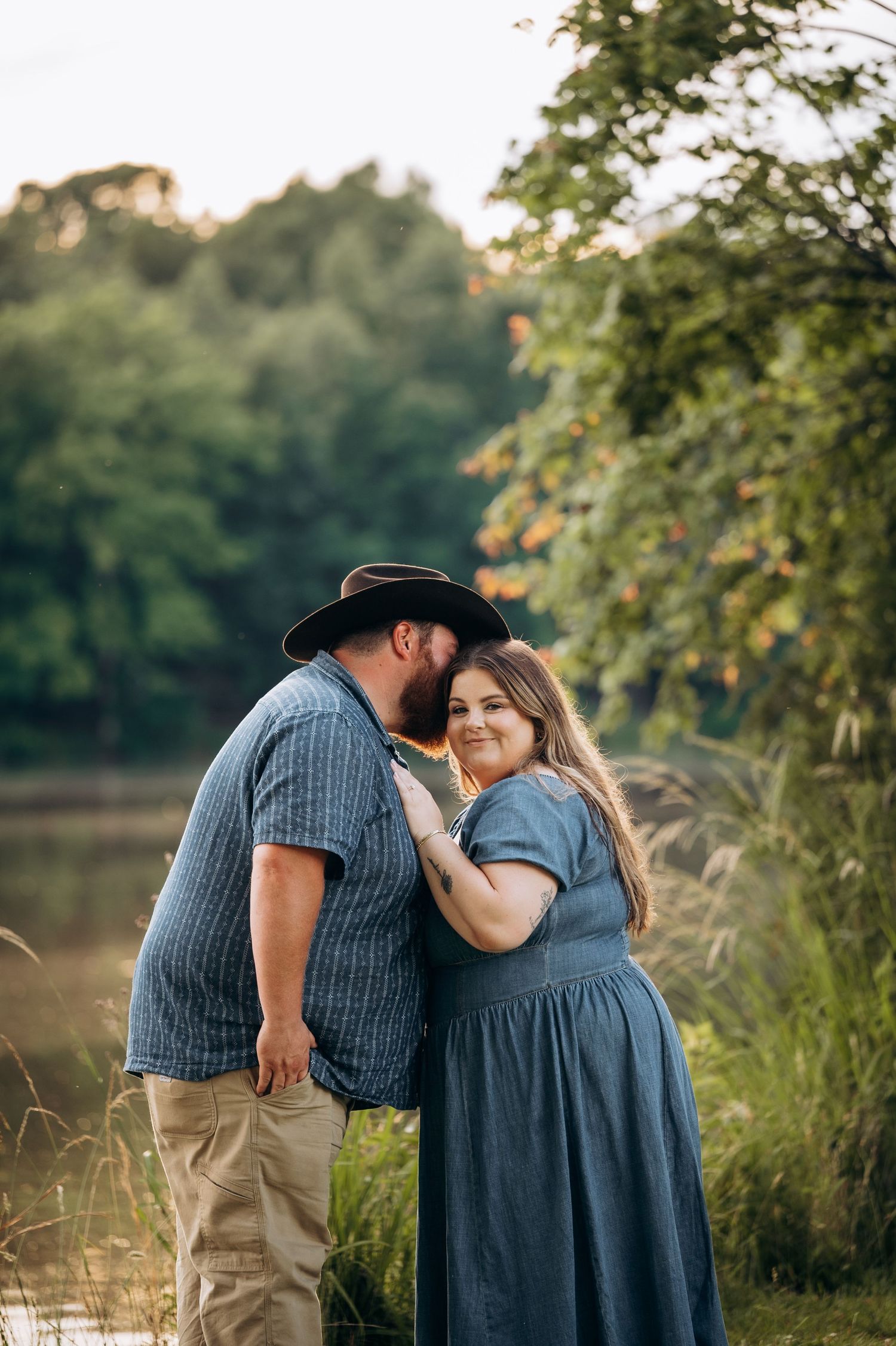 Man in cowboy hat kissing woman in blue dress near a sunlit lake surrounded by green trees.