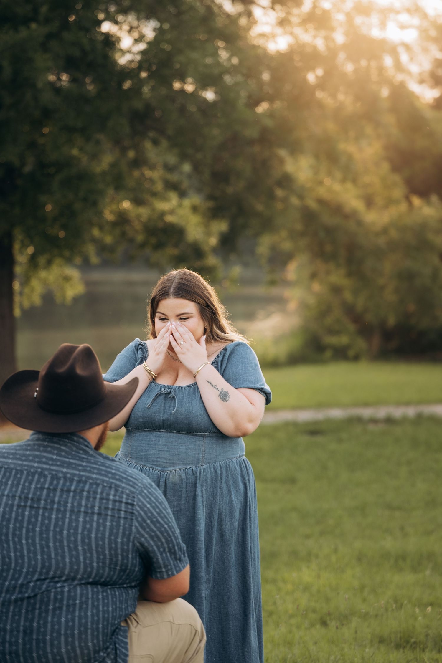 Emotional woman in blue dress covers her mouth in surprise during an outdoor proposal at sunset.