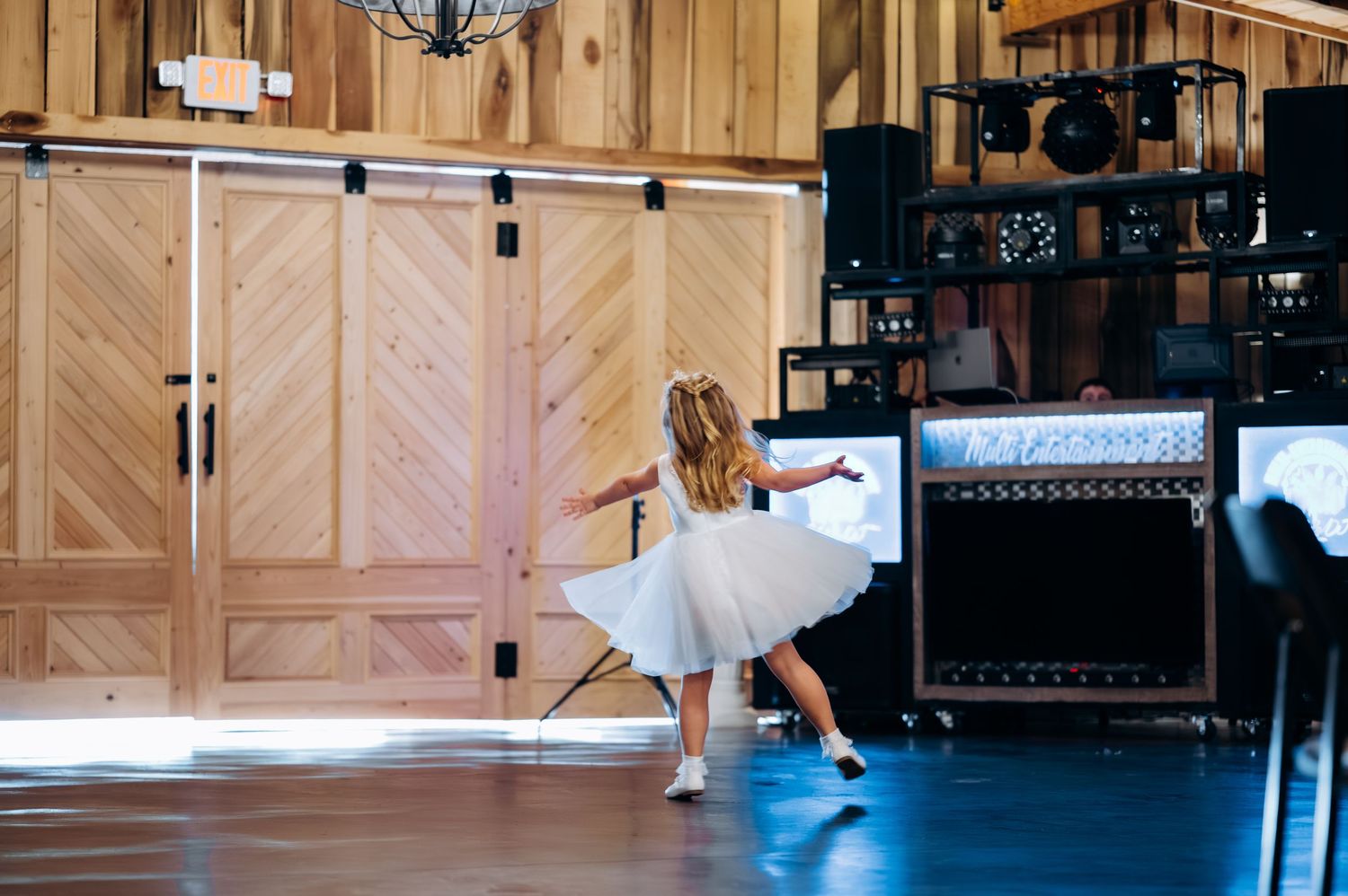 Young flower girl in white dress twirling on dance floor in front of rustic wooden barn doors at wedding reception.