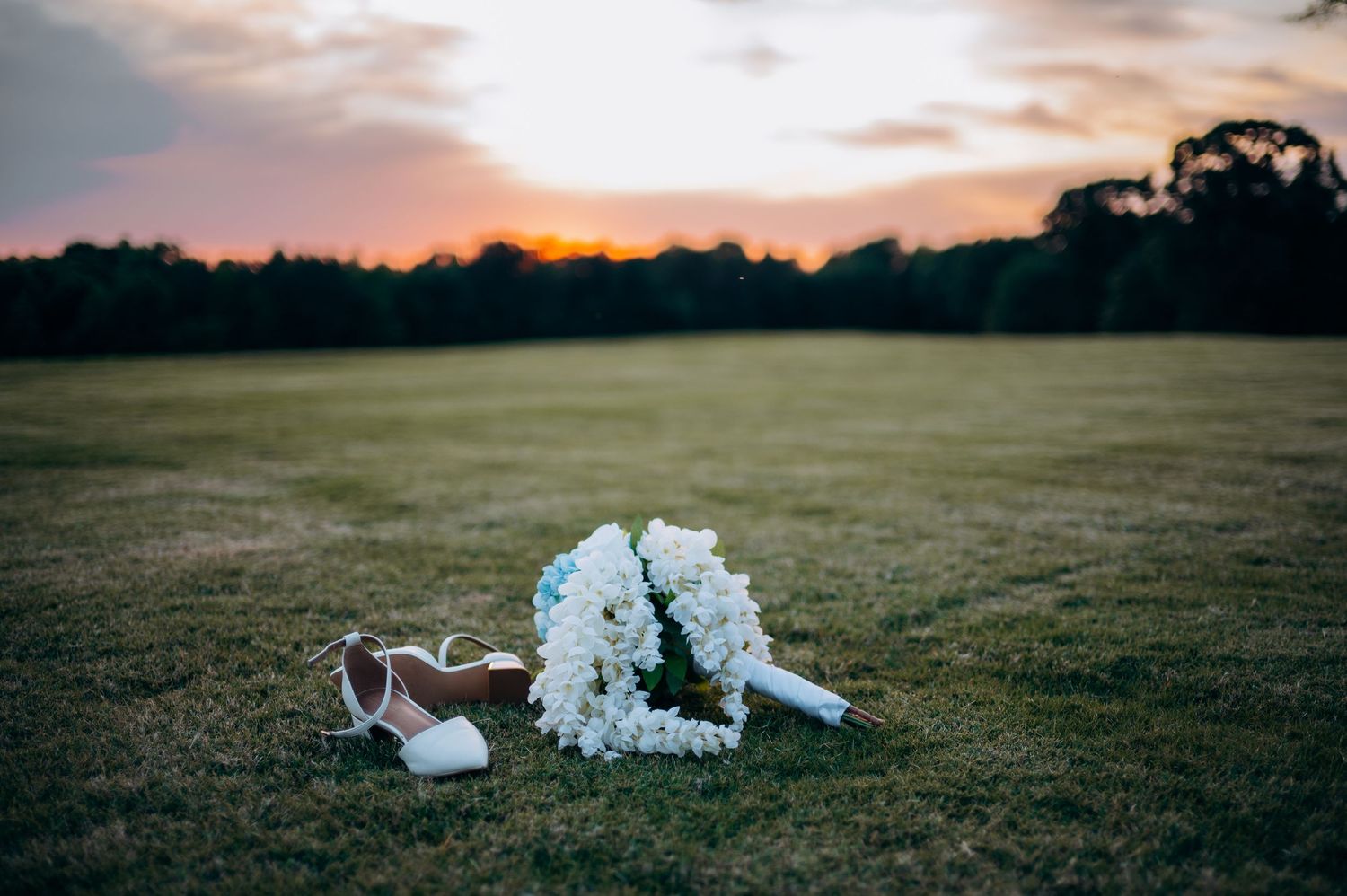White bridal bouquet and wedding heels resting on green grass field at sunset with warm golden sky backdrop.