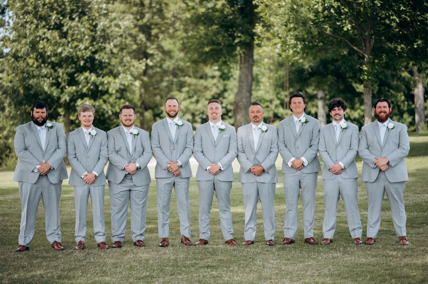Ten groomsmen in matching light gray suits stand in a line outdoors on a green lawn for a wedding photo.