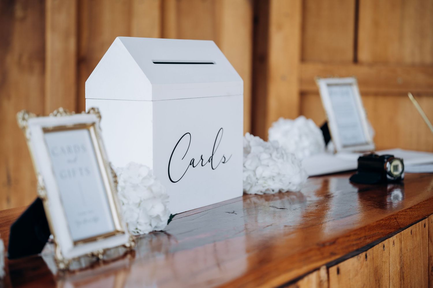 White wedding card box with elegant script lettering on a wooden table, flanked by white hydrangeas and framed signs.