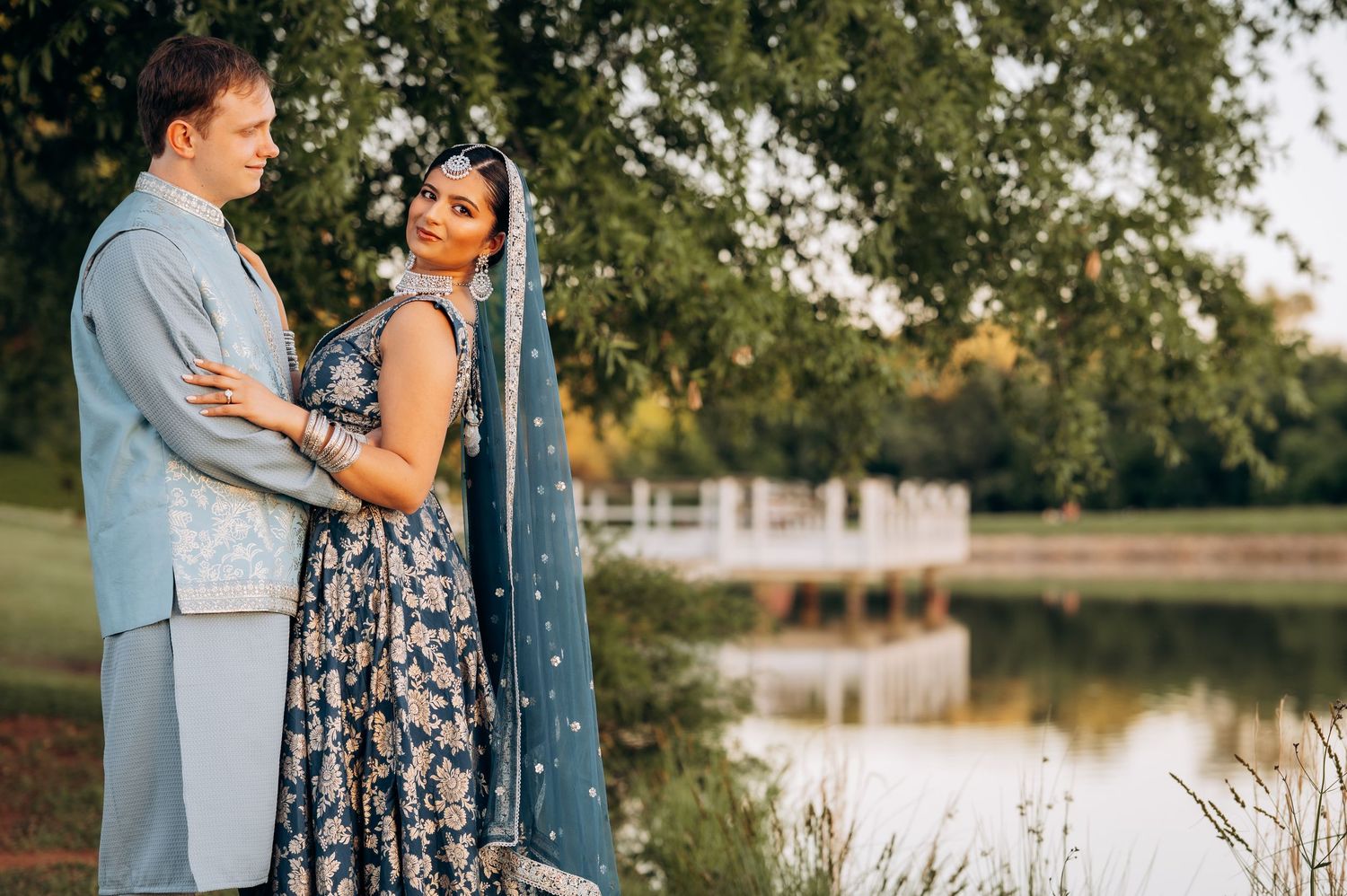 Couple in coordinating blue outfits posing near a serene pond, woman wearing elegant Indian attire with embroidered dupatta.