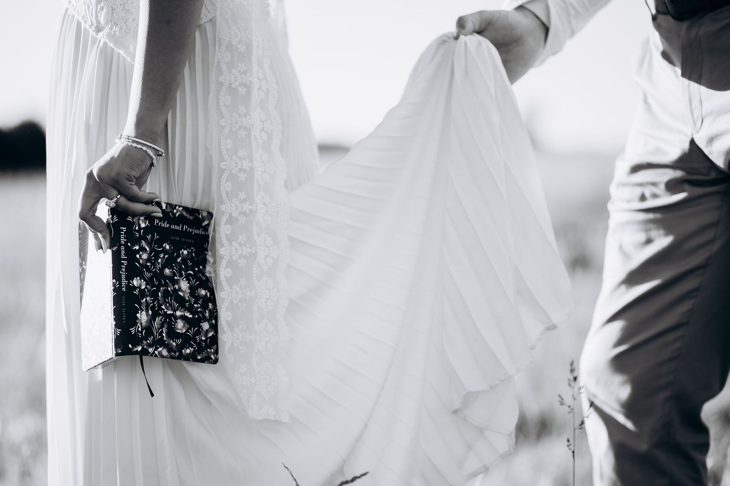 Black and white photo of a bride in a flowing white gown holding a clutch, with a man lifting her dress train.