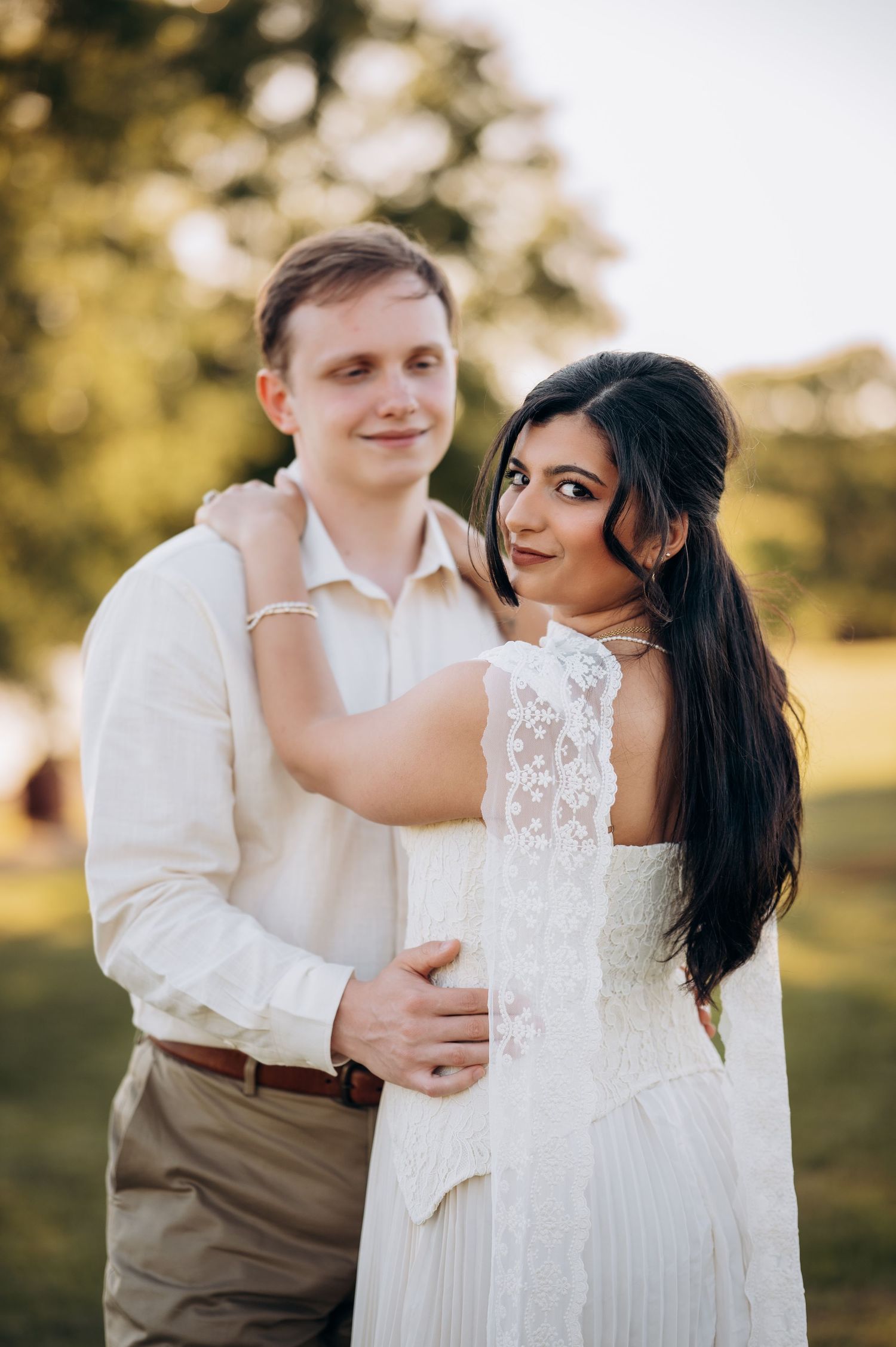 Couple in wedding attire embracing outdoors, bride in white lace dress looking back, groom in khaki suit smiling.